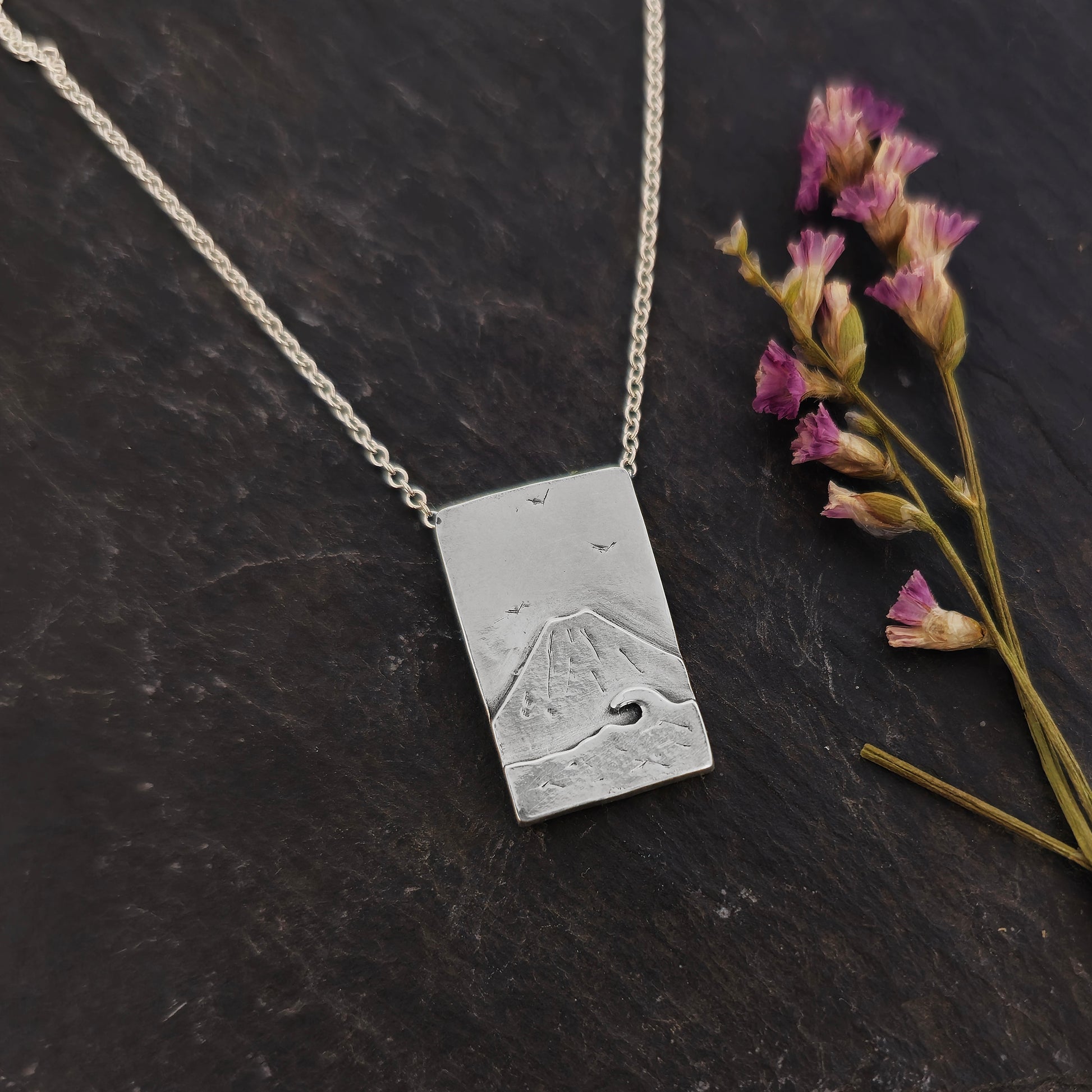 A rectangular silver pendant with a mountain scene featuring Mount Fuji behind a sea with a wave and birds in the sky. On a silver chain with invisible bail. Mijoux Creations. Shown on slate with flowers.