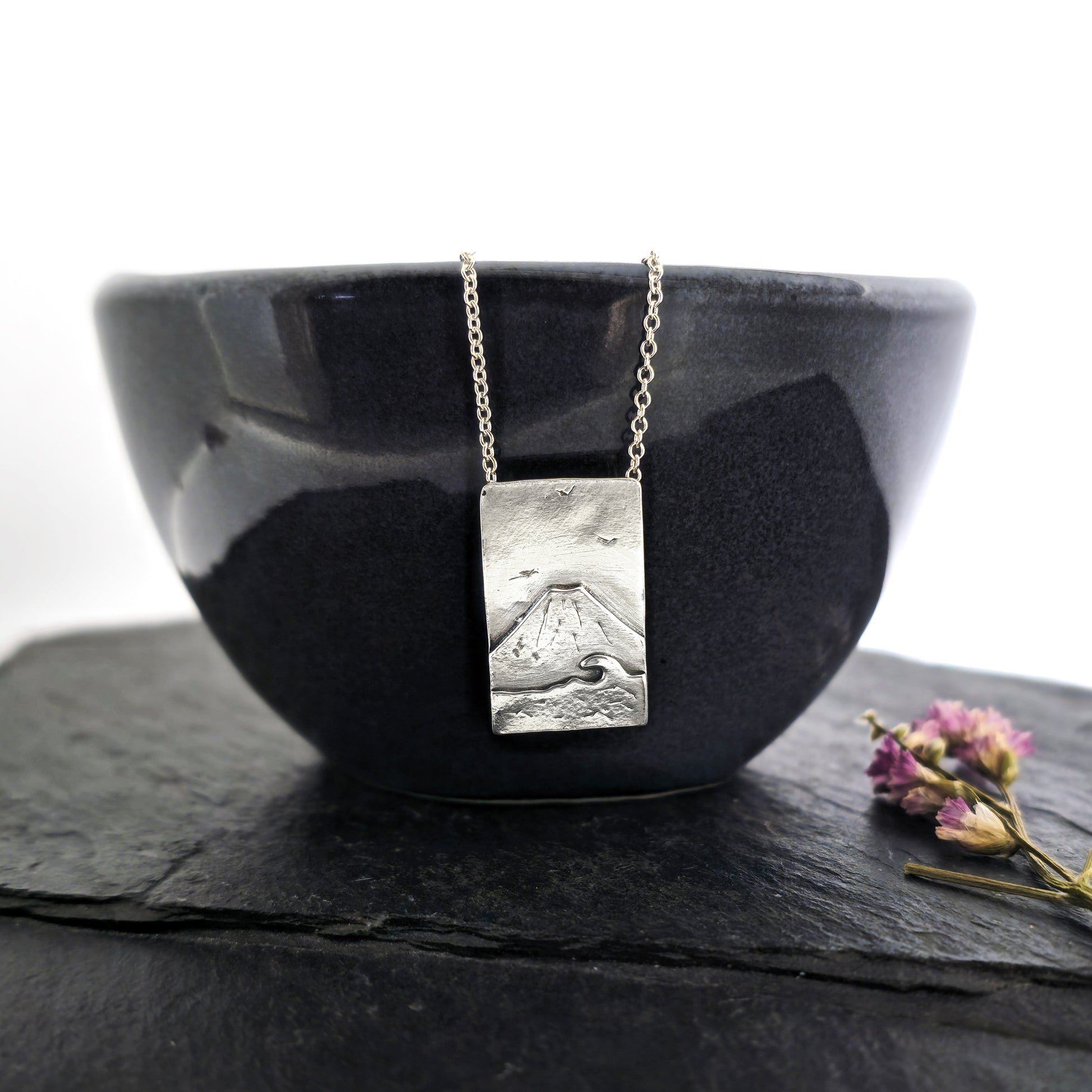 A rectangular silver pendant with a mountain scene featuring Mount Fuji behind a sea with a wave and birds in the sky. On a silver chain with invisible bail. Mijoux Creations. Shown on a black bowl with flowers.