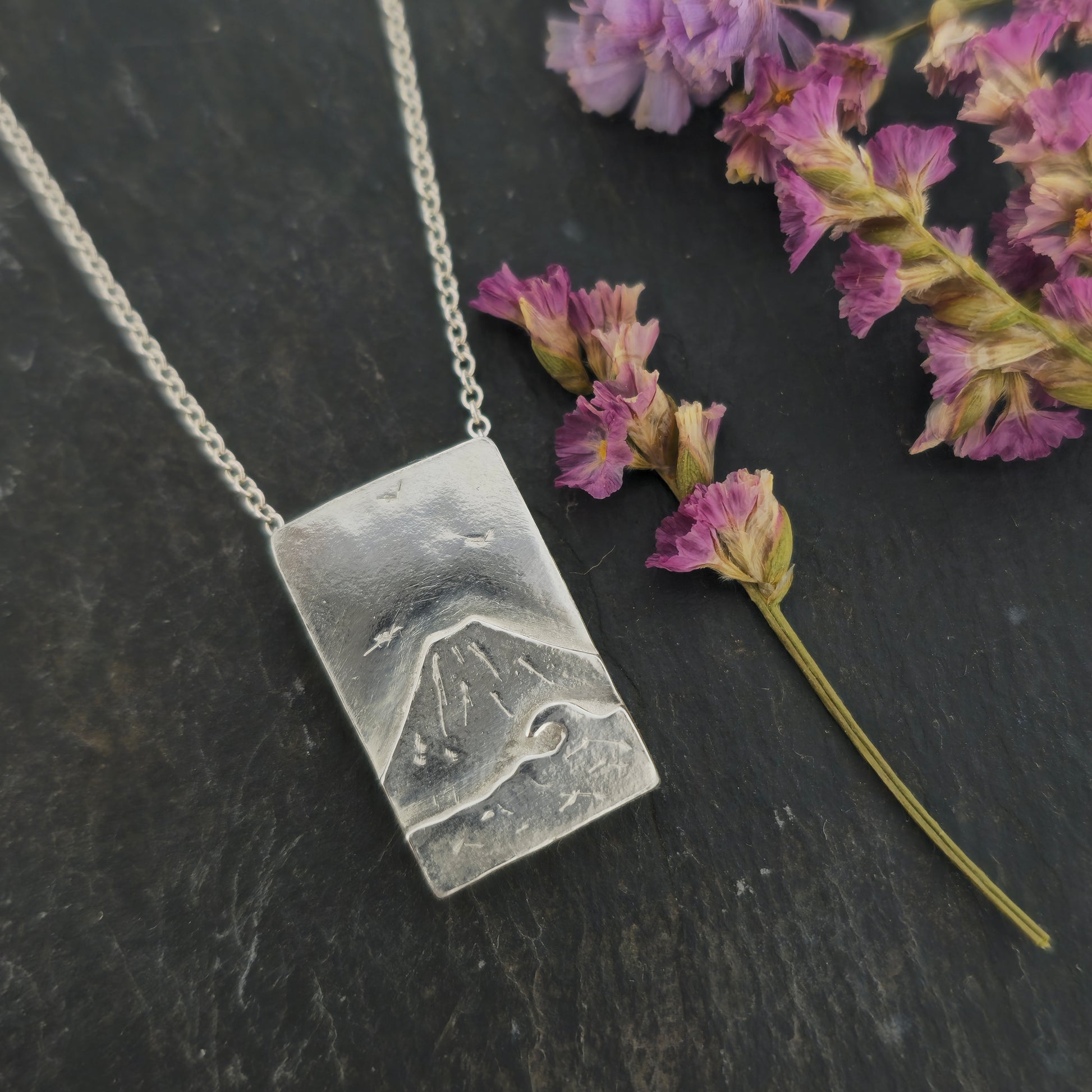 A rectangular silver pendant with a mountain scene featuring Mount Fuji behind a sea with a wave and birds in the sky. On a silver chain with invisible bail. Shown on slate with flowers.