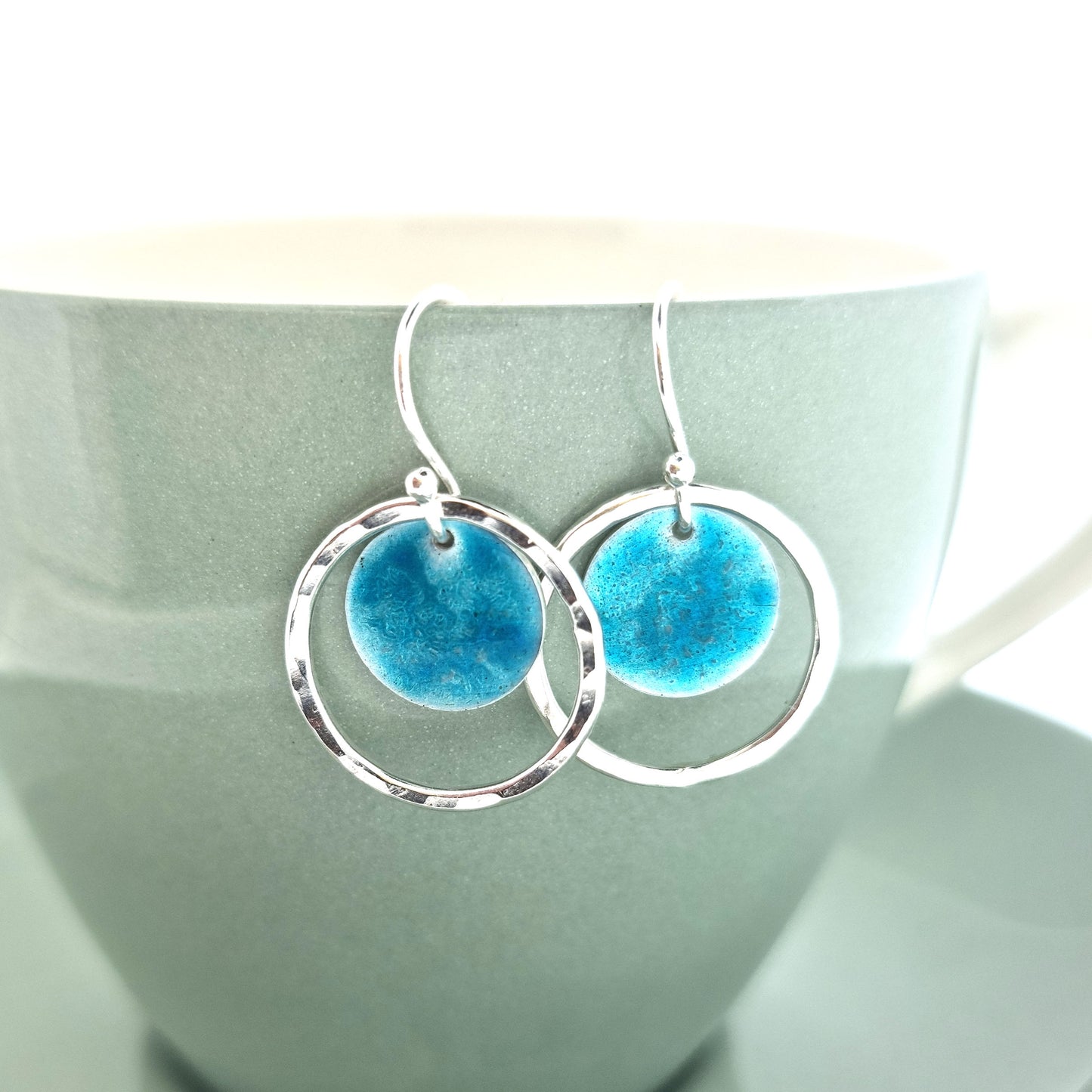 Silver drop earrings with a central silver disc covered with a turquoise enamel pattern and a surrounding hammered silver circle. Shown on cup.