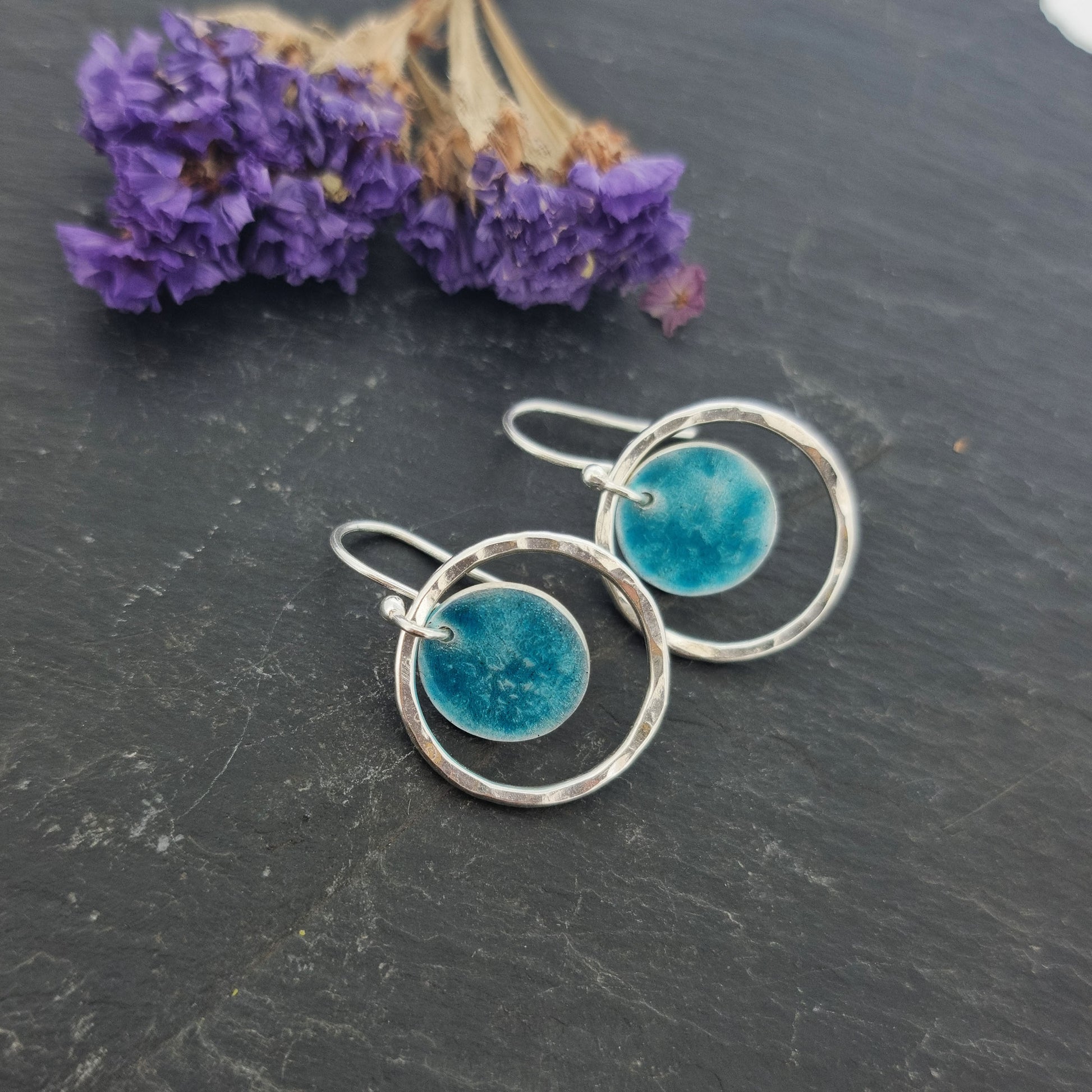 Silver drop earrings with a central silver disc covered with a turquoise enamel pattern and a surrounding hammered silver circle. Pictured on slate with flowers.