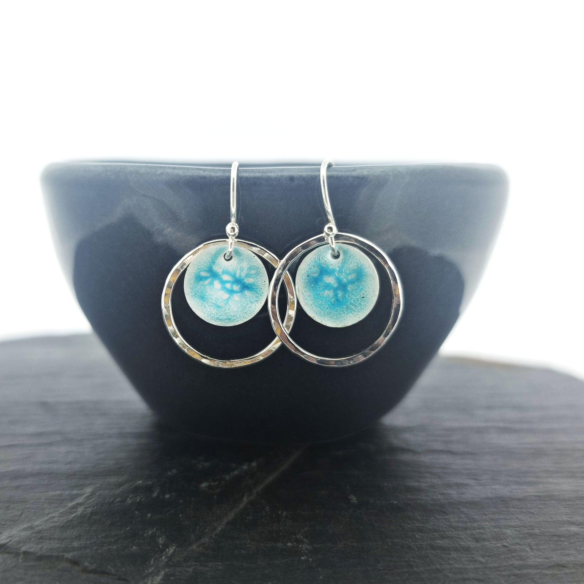 Silver drop earrings with a central silver disc covered with a turquoise enamel pattern and a surrounding hammered silver circle. Pictured on a black bowl.