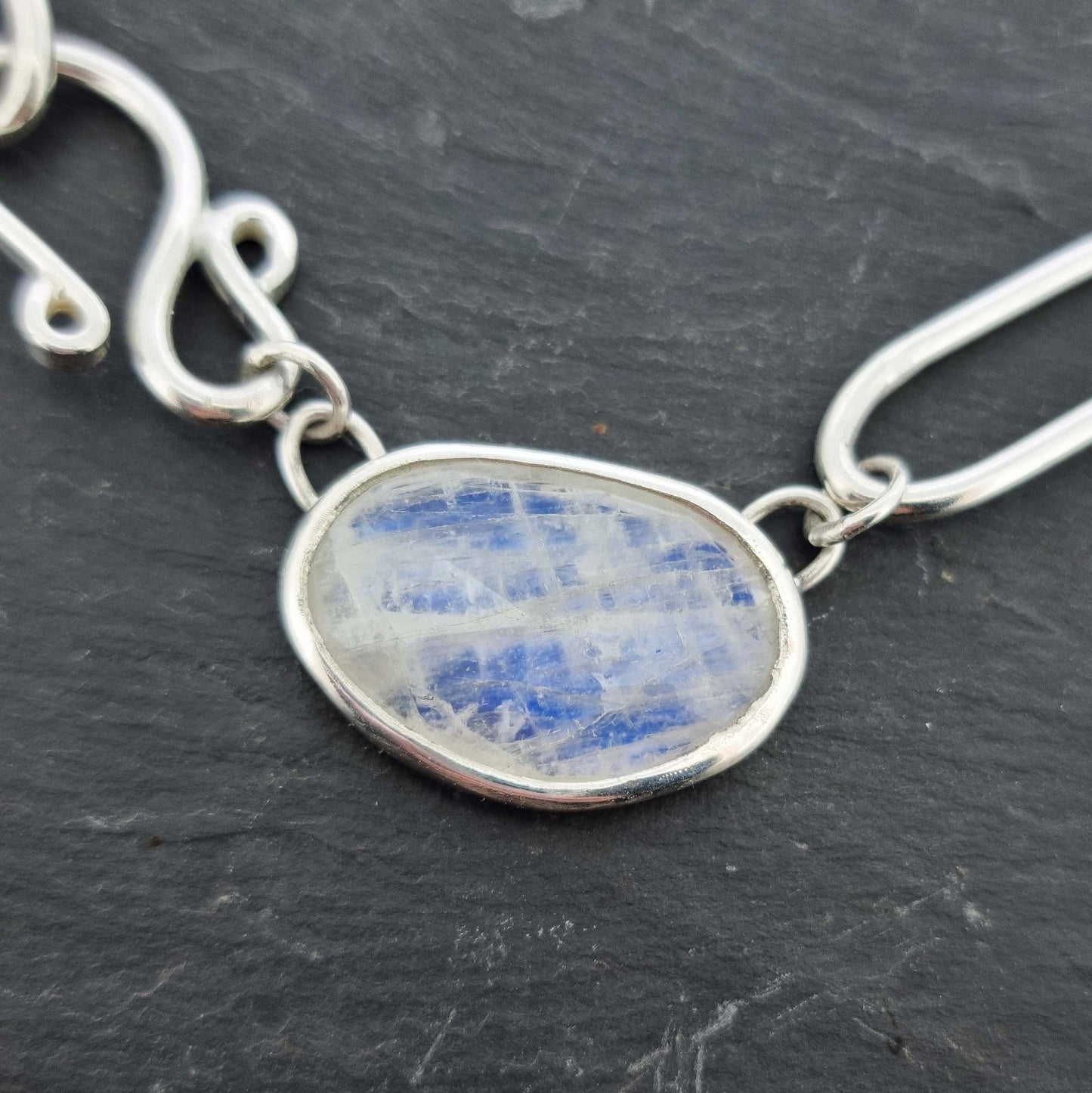 A silver chain necklace with open oblong links of different sizes, an s-clasp and a moonstone slice in a bezel setting.