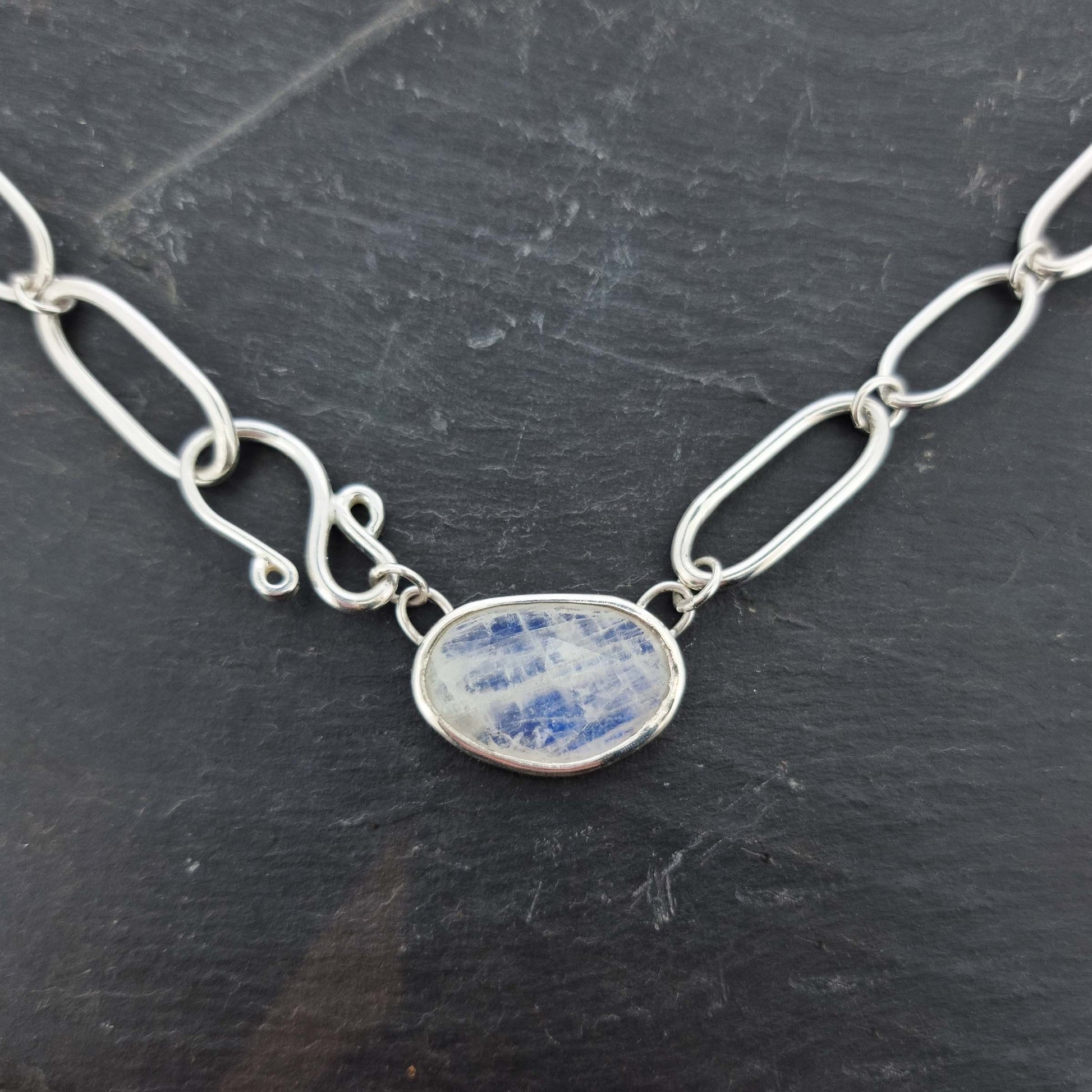 A silver chain necklace with open oblong links of different sizes, an s-clasp and a moonstone slice in a bezel setting.