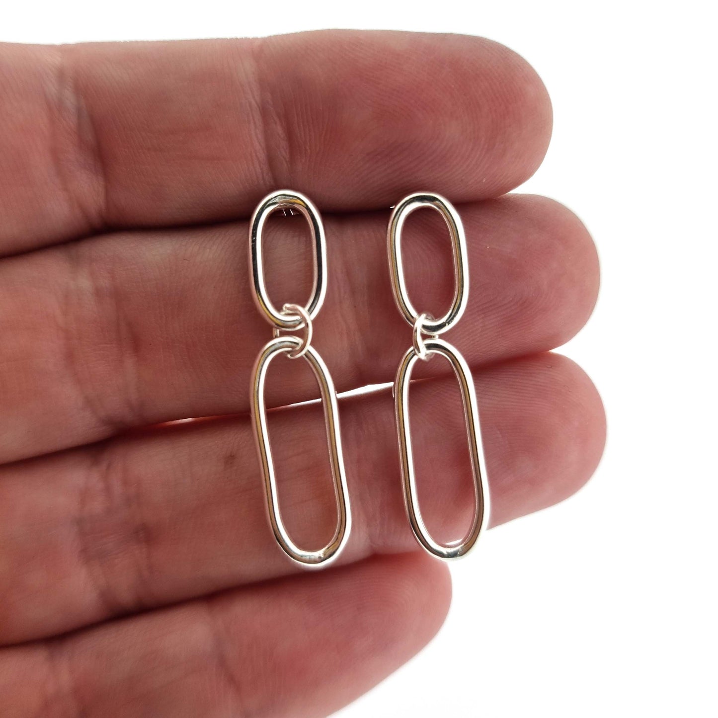 Silver drop earrings featuring two open oblong links of different sizes. Being held by a hand.