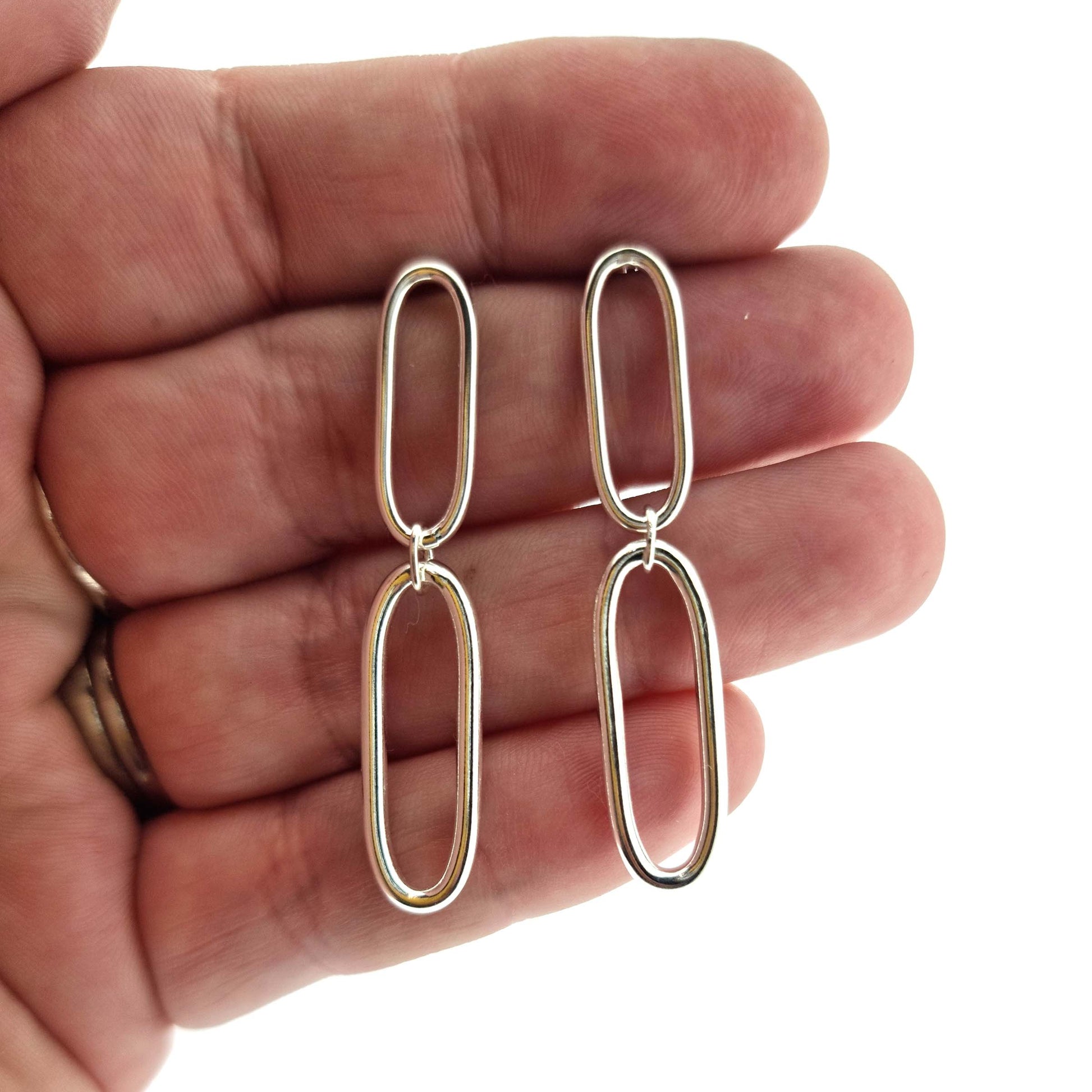 Silver drop earrings featuring 2 open oblong oval shapes attached to each other in a long drop. Shown held in a hand.