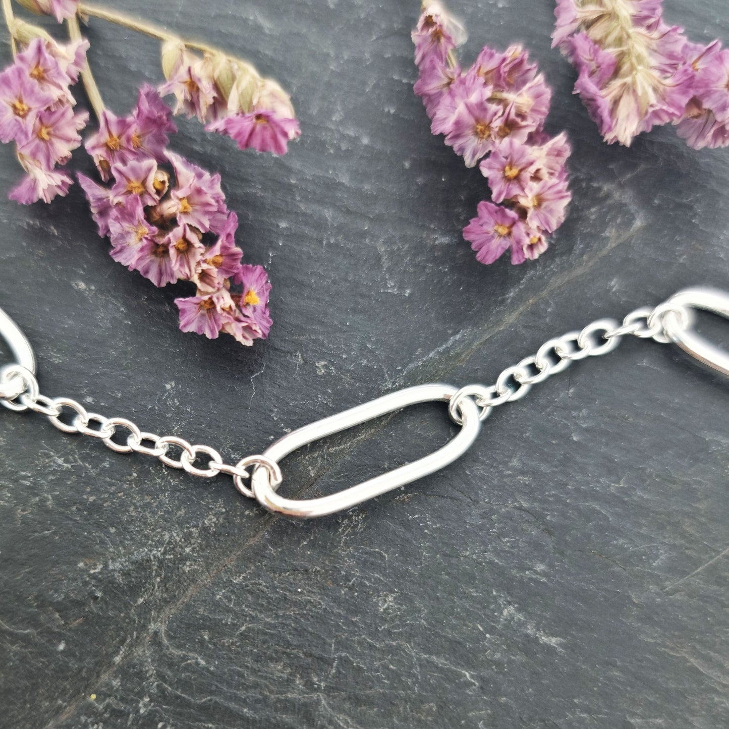 A close up of a silver bracelet with oblong oval links set between round linked chain. Pictured on slate with flowers.