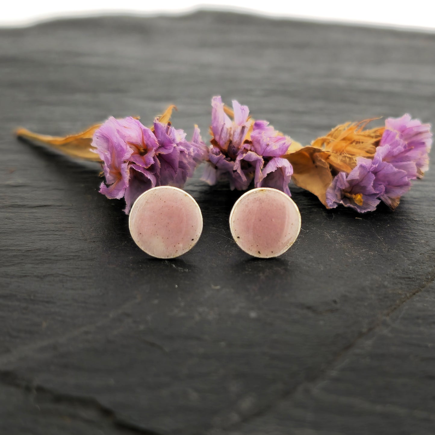 A pair of round silver stud earrings with a pale pink enamel finish. Pictured on slate with flowers.