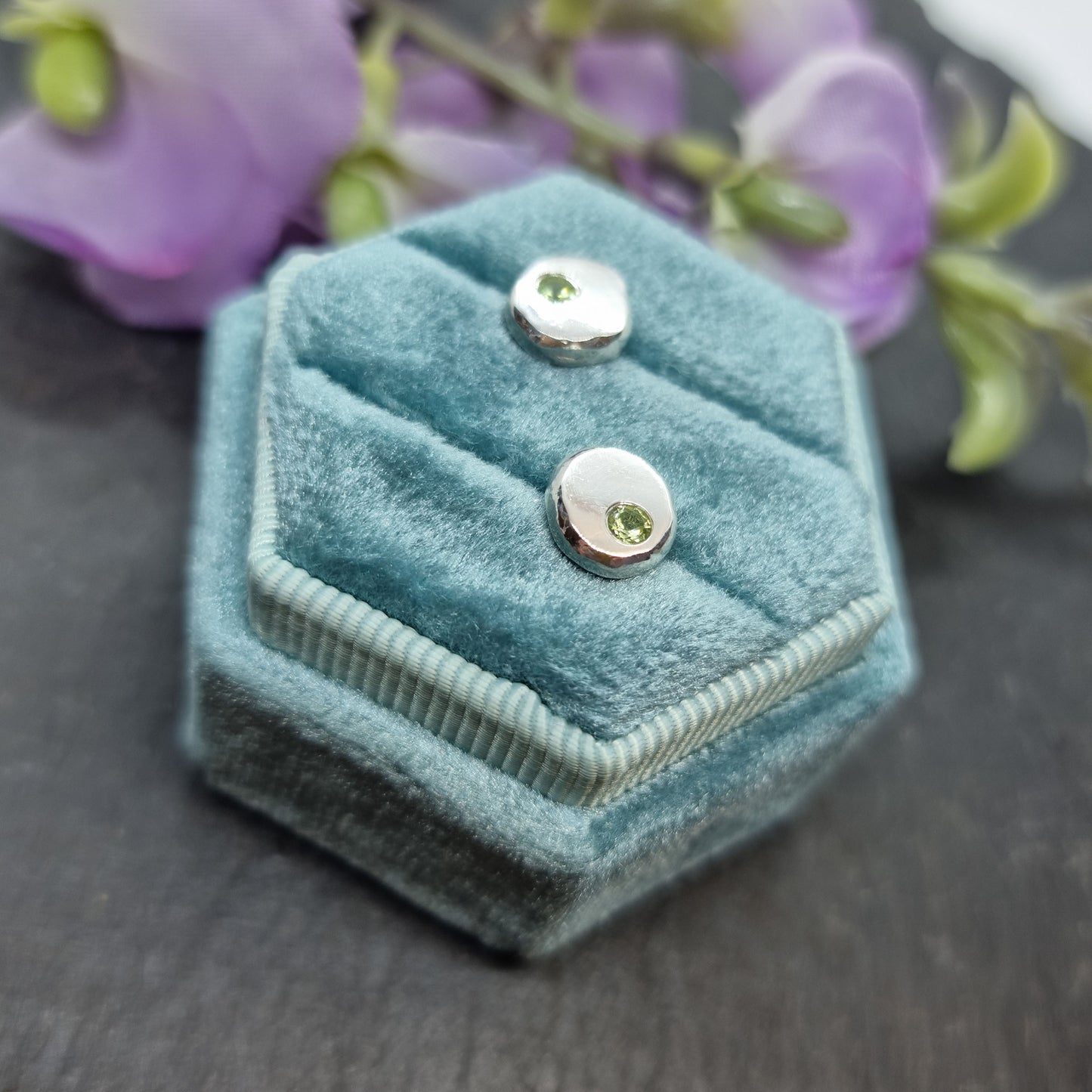 Silver round flat 'pebble' stud earrings set with pale green peridot gemstones. In jewellery box with flowers.