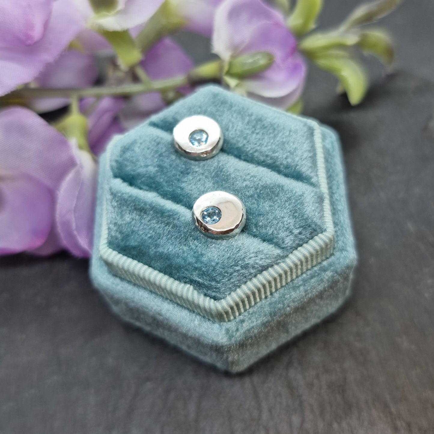 Silver round stud earrings with round blue topaz gemstones flush set into them. Shown in jewellery box with flowers.