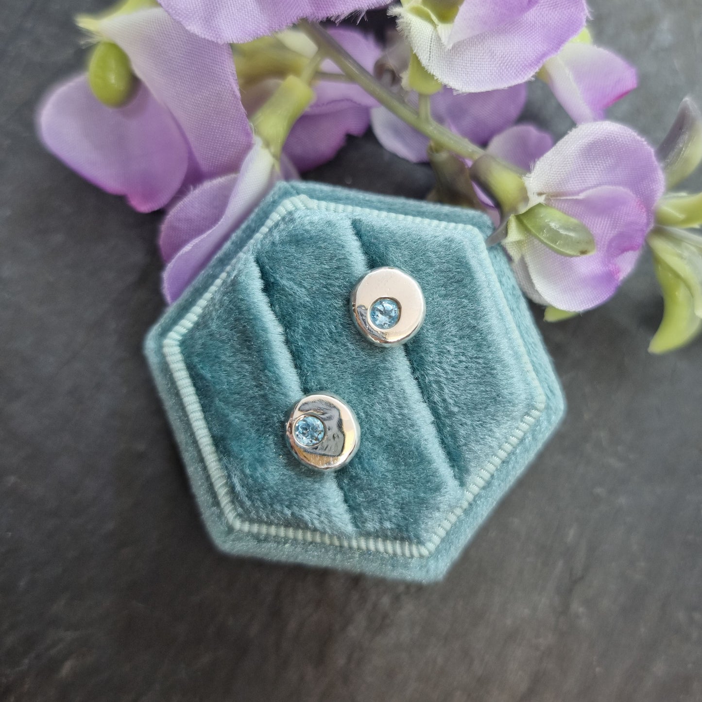 Silver round stud earrings with round blue topaz gemstones flush set into them. Shown in jewellery box with flowers.