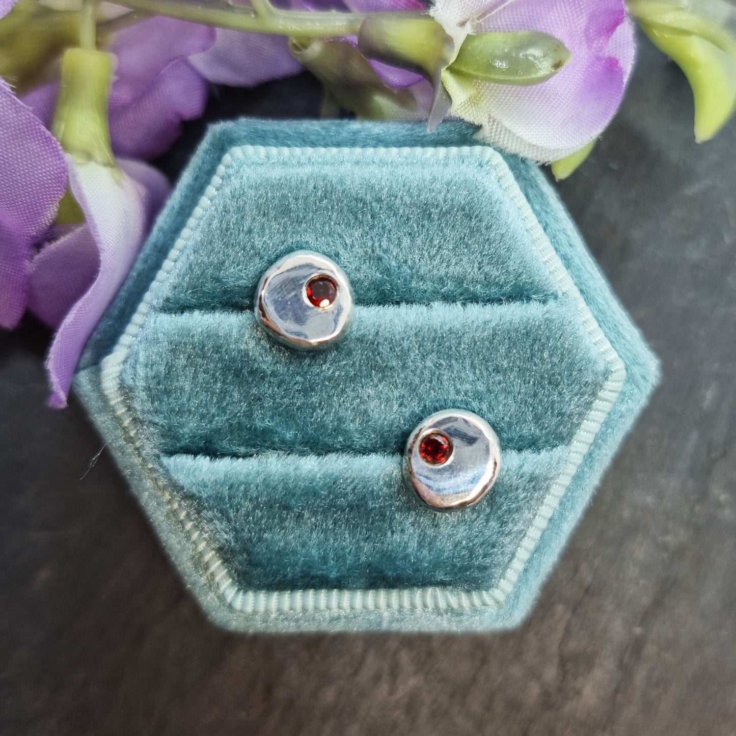 Round silver stud earrings with red garnet gemstones set off-centre. Pictured in a jewellery box with flowers.