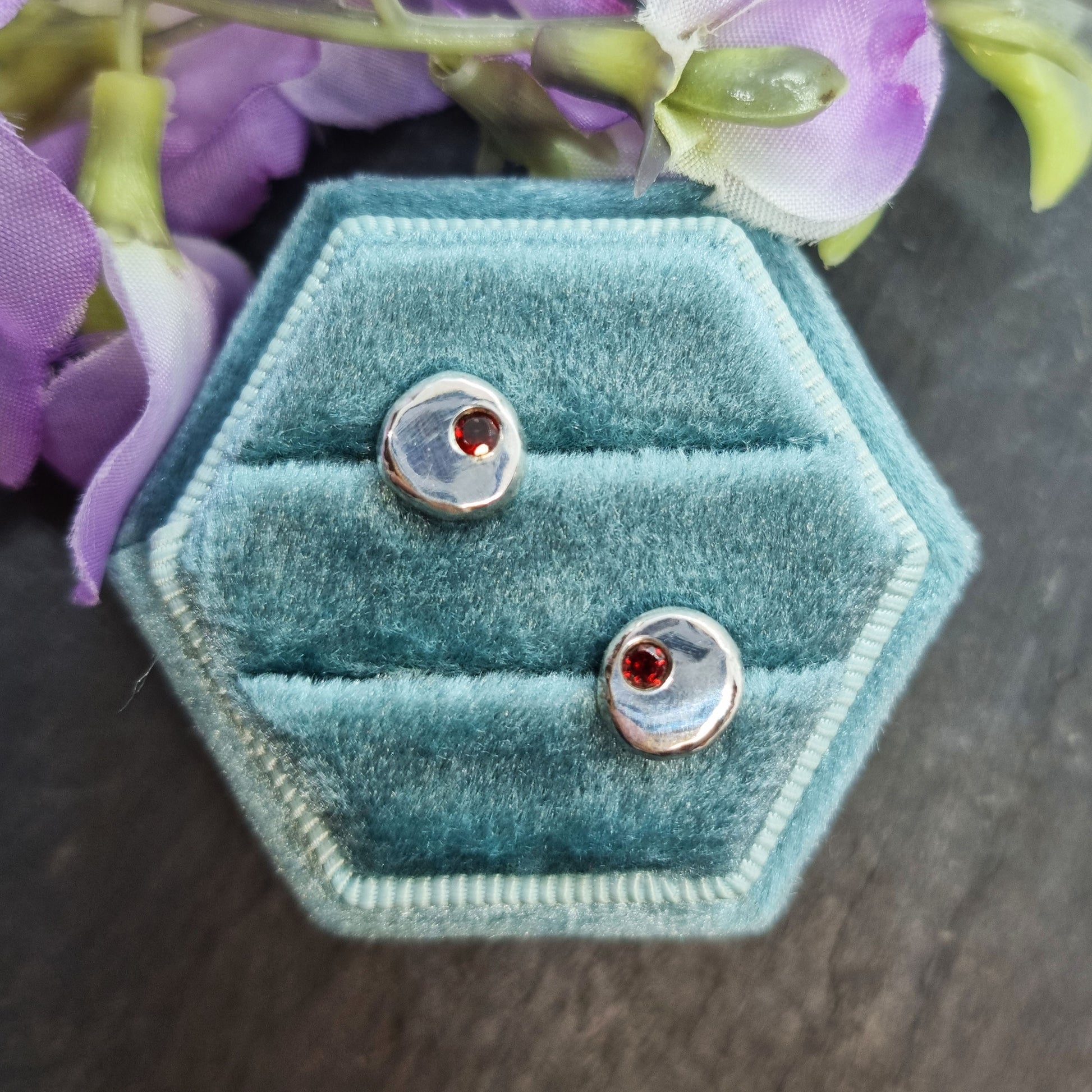 Round silver stud earrings with red garnet gemstones set off-centre. Pictured in a jewellery box with flowers.