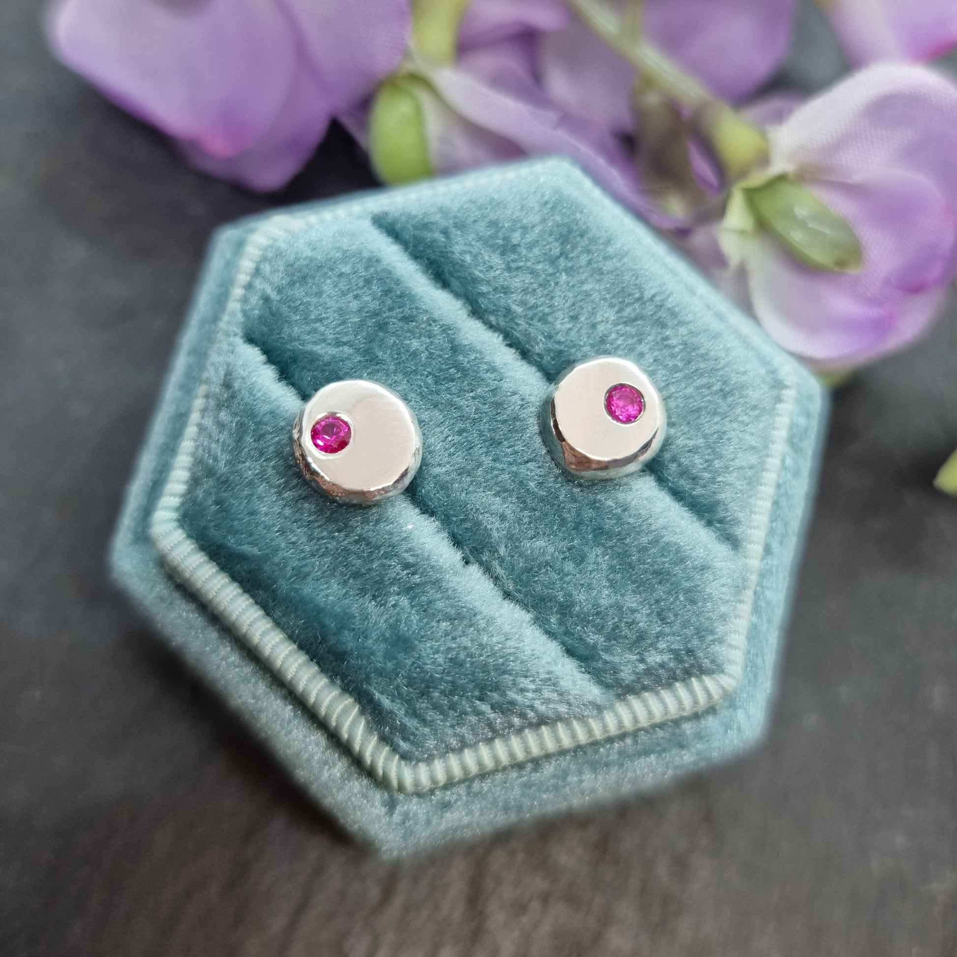 Round silver pebble stud earrings with red rubies flush set off-centre. Pictured in a jewellery box with flowers.