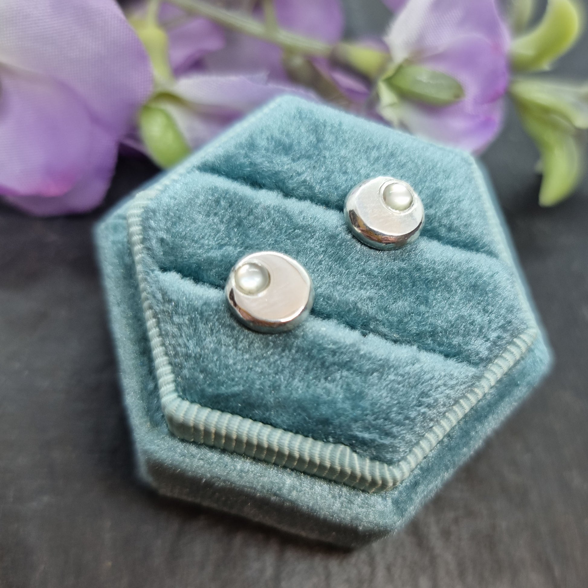 Round silver pebble stud earrings with mother of pearl gemstones flush set off-centre. Pictured in a jewellery box with flowers.