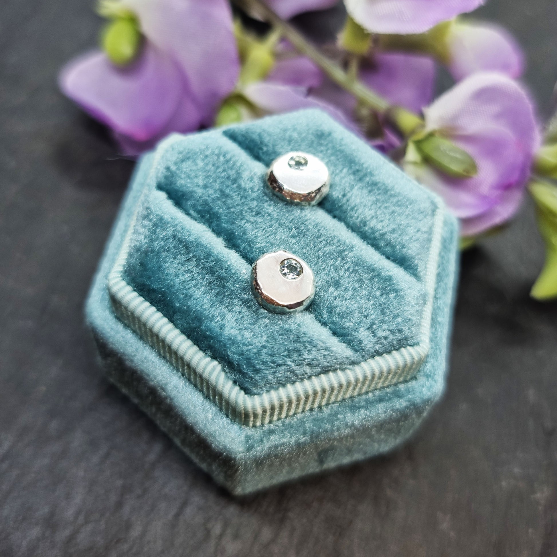 Round silver pebble stud earrings with pale blue aquamarine gemstones set off-centre. Pictured in a jewellery box with flowers.