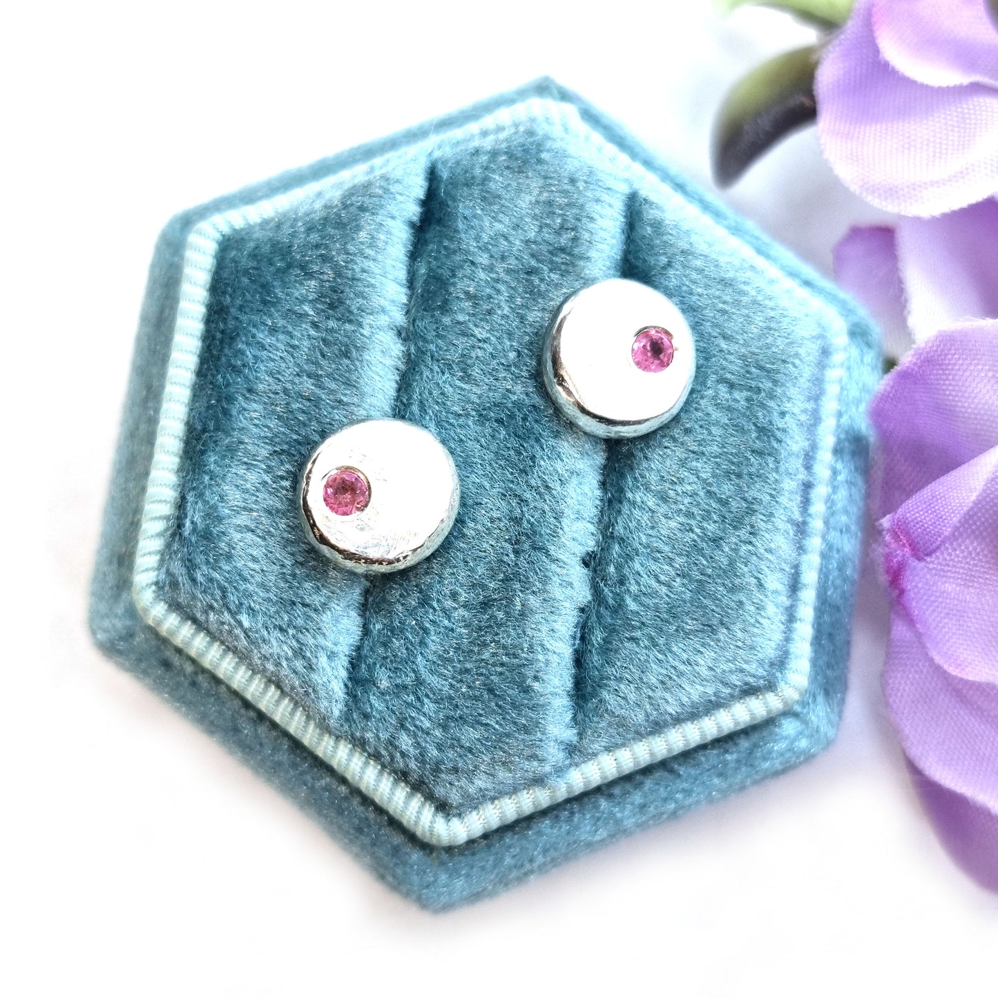 Round silver pebble stud earrings with pink tourmaline gemstones flush set off-centre. Pictured in a jewellery box with flowers.