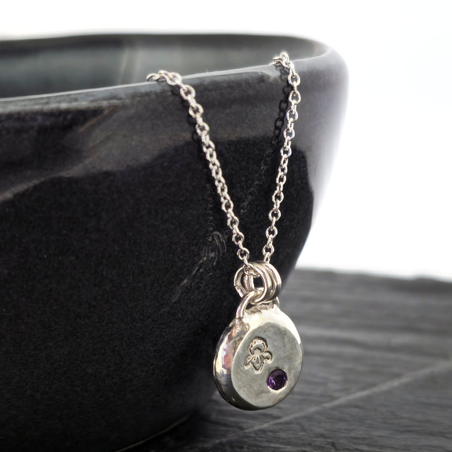 A handmade round silver pendant with a violet flower (February birth flower) engraved on it and a round purple amethyst (February birthstone) flush set into it. On a silver chain. On a black bowl. Mijoux Jewellery.