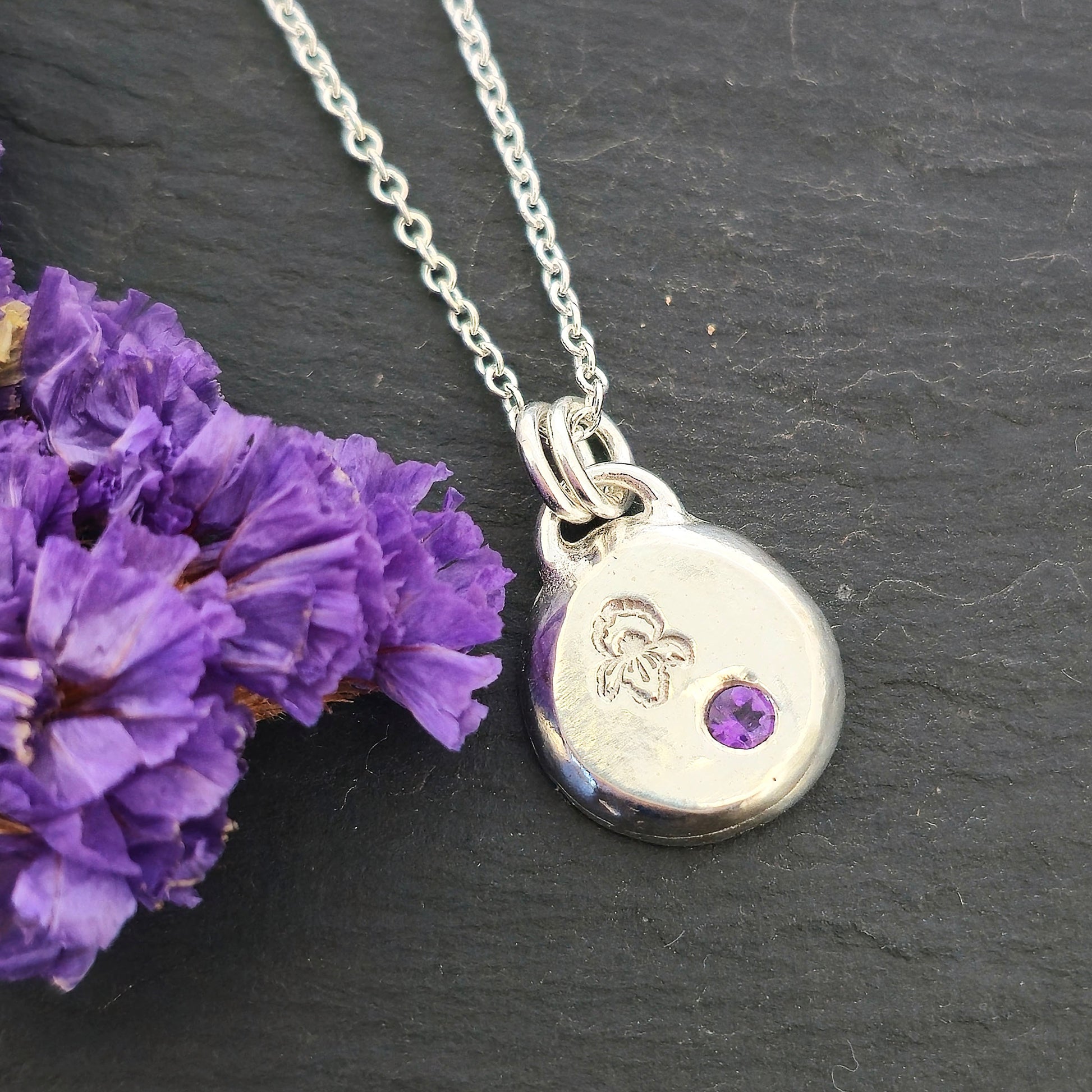 A handmade round silver pendant with a violet flower (February birth flower) engraved on it and a round purple amethyst (February birthstone) flush set into it. On a silver chain. On a black background with purple flowers. Mijoux Jewellery.