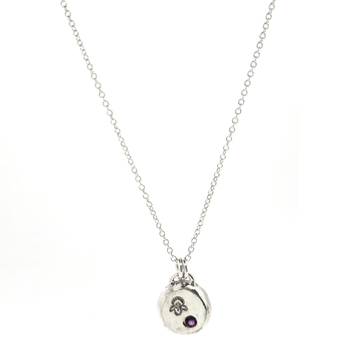 A handmade round silver pendant with a violet flower (February birth flower) engraved on it and a round purple amethyst (February birthstone) flush set into it. On a silver chain. On a white background. Mijoux Jewellery.