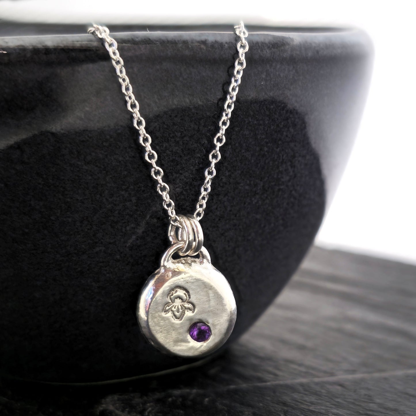 A handmade round silver pendant with a violet flower (February birth flower) engraved on it and a round purple amethyst (February birthstone) flush set into it. On a silver chain. On a black bowl. Mijoux Jewellery.