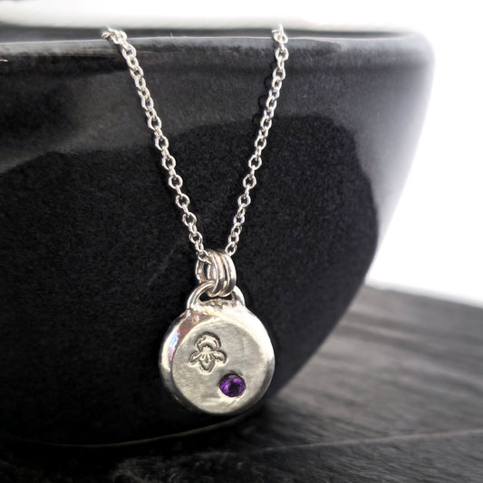 A handmade round silver pendant with a violet flower (February birth flower) engraved on it and a round purple amethyst (February birthstone) flush set into it. On a silver chain. On a black bowl. Mijoux Jewellery.