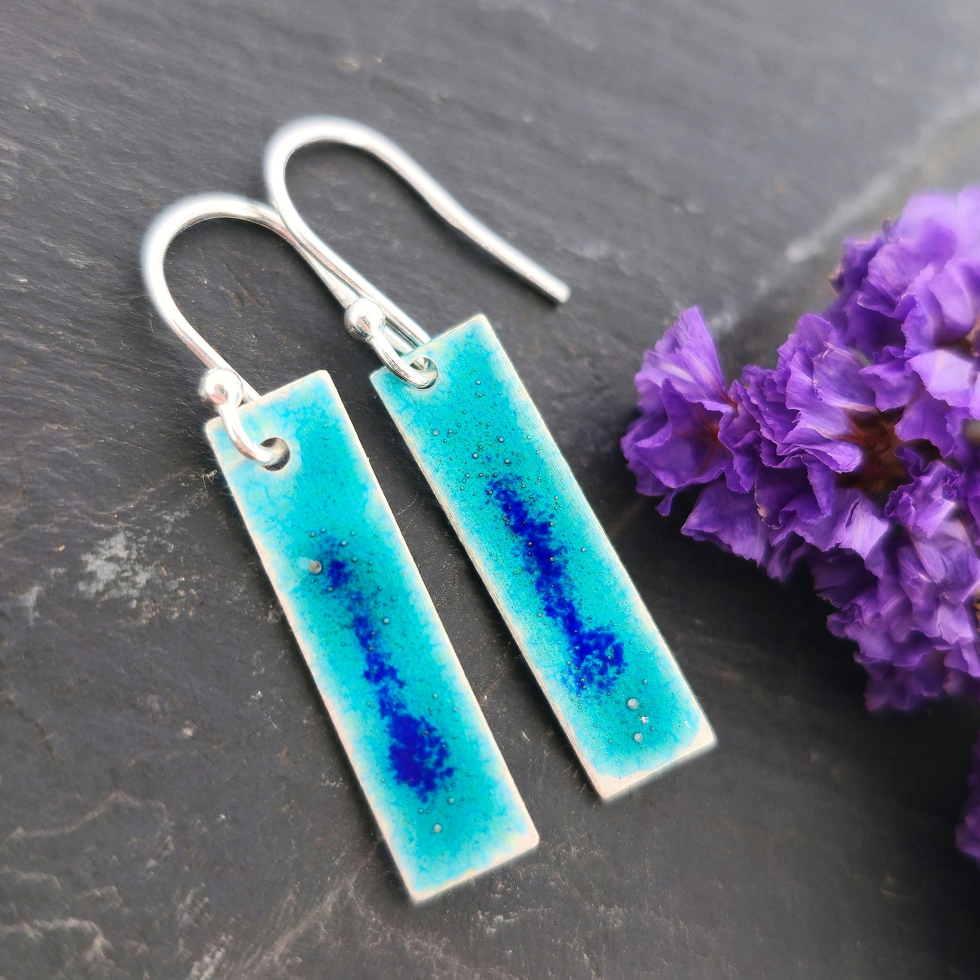 Rectangular silver drop earrings with a turquoise enamel background and a speckled line of dark blue enamel in the middle. On silver ear hooks. Shown on black background with purple flowers. Mijoux Jewellery.