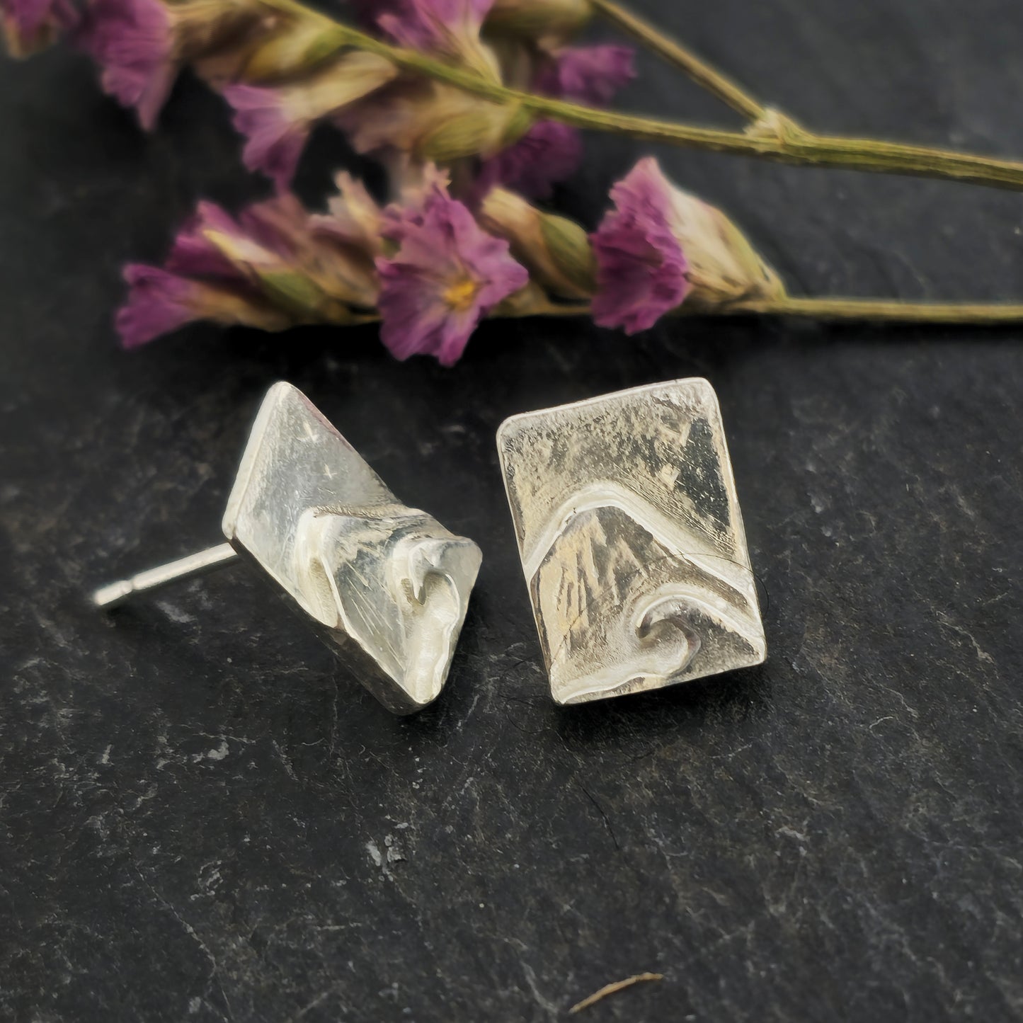 Rectangular silver stud earrings with a mountain scene featuring a sea with a large wave in front of Mount Fuji and birds flying in the sky. Pictured on slate with flowers.