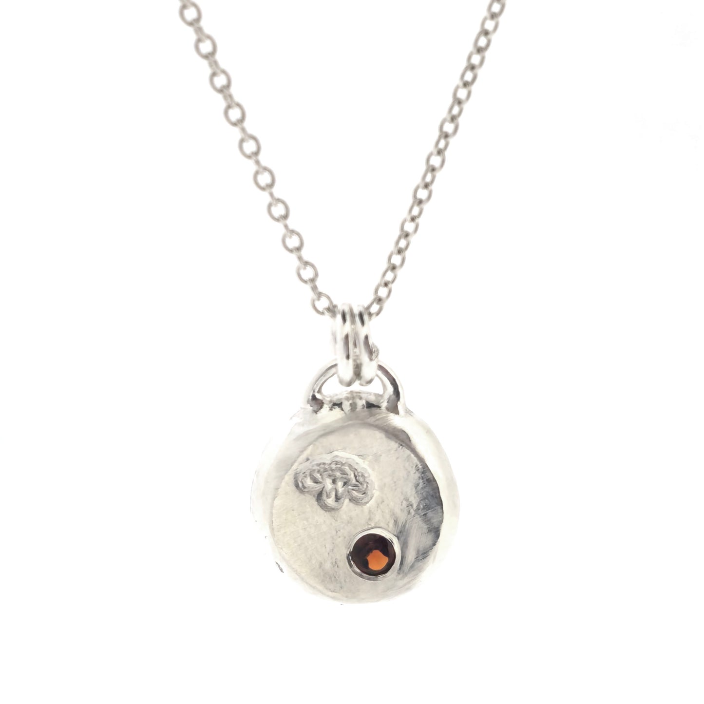 A round silver flat pebble pendant with a carnation flower (January's birth flower) engraved on it and a red garnet gemstone (January's birthstone) flush set into it. On a silver chain. On a white background.