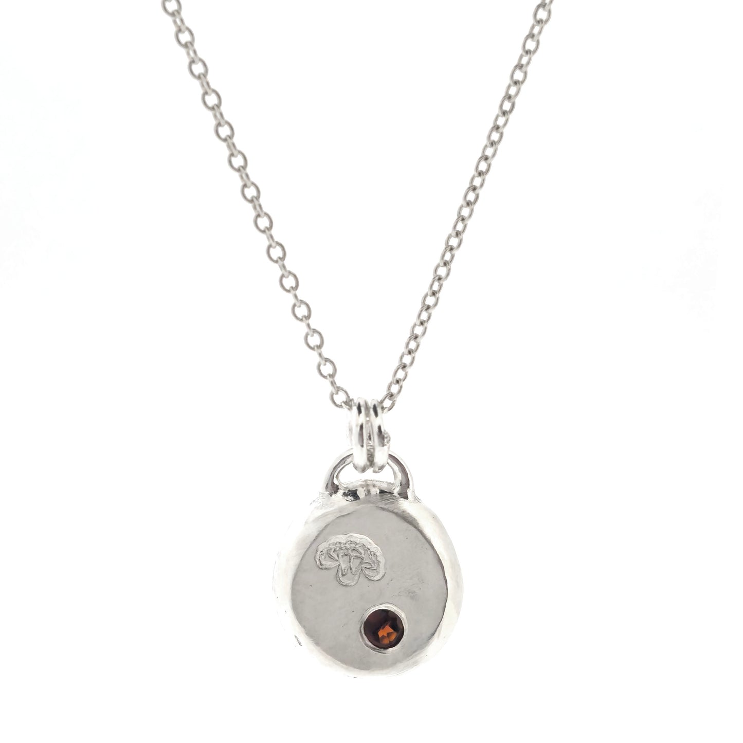 A round silver flat pebble pendant with a carnation flower (January's birth flower) engraved on it and a red garnet gemstone (January's birthstone) flush set into it. On a silver chain. On a white background.