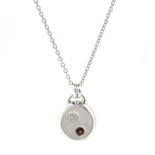A round silver flat pebble pendant with a carnation flower (January's birth flower) engraved on it and a red garnet gemstone (January's birthstone) flush set into it. On a silver chain. On a white background.