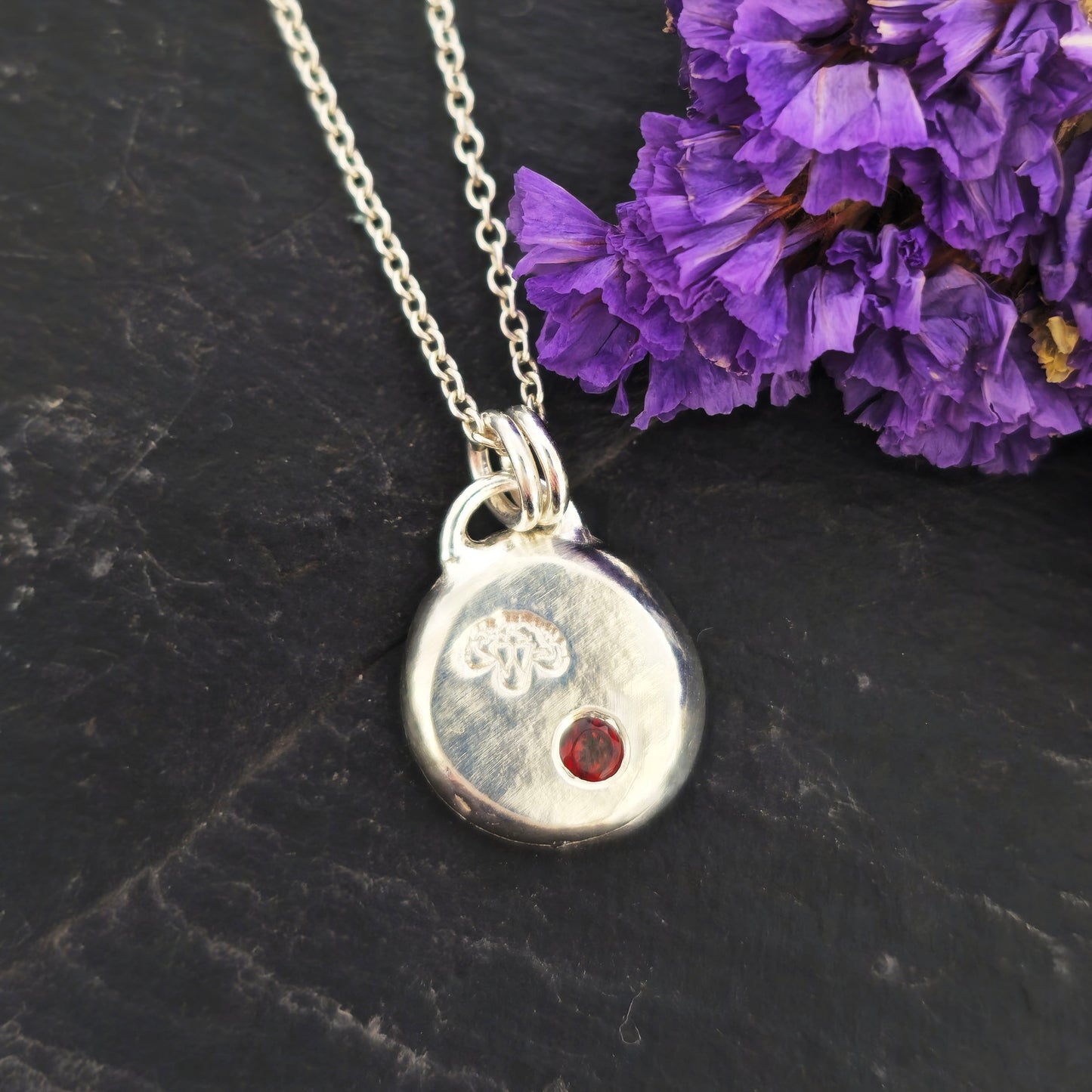 A round silver flat pebble pendant with a carnation flower (January's birth flower) engraved on it and a red garnet gemstone (January's birthstone) flush set into it. On a silver chain. On a black background with purple flowers. Mijoux Jewellery.