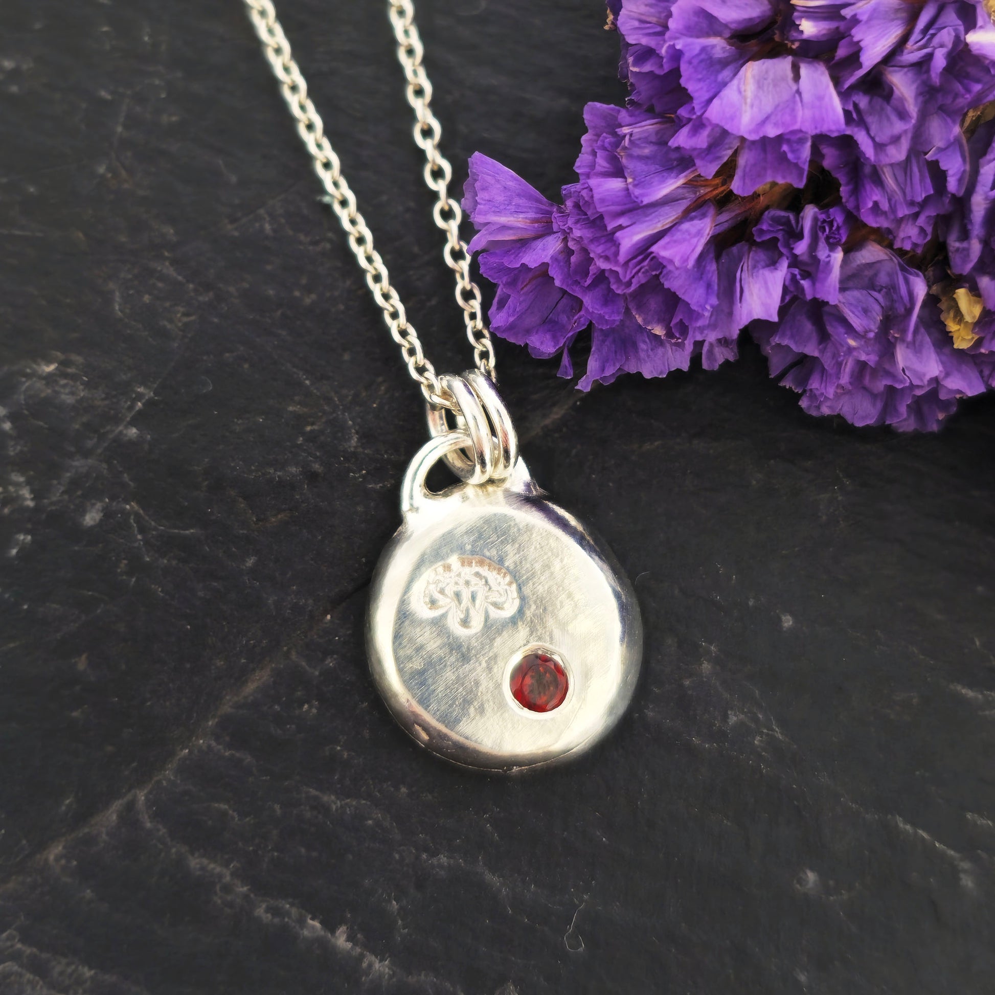 A round silver flat pebble pendant with a carnation flower (January's birth flower) engraved on it and a red garnet gemstone (January's birthstone) flush set into it. On a silver chain. On a black background with purple flowers. Mijoux Jewellery.