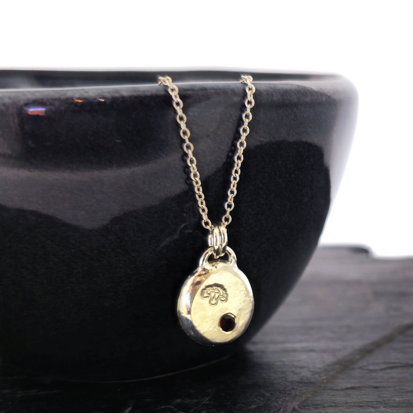 A round silver flat pebble pendant with a carnation flower (January's birth flower) engraved on it and a red garnet gemstone (January's birthstone) flush set into it. On a silver chain. On a black bowl. Mijoux Jewellery.