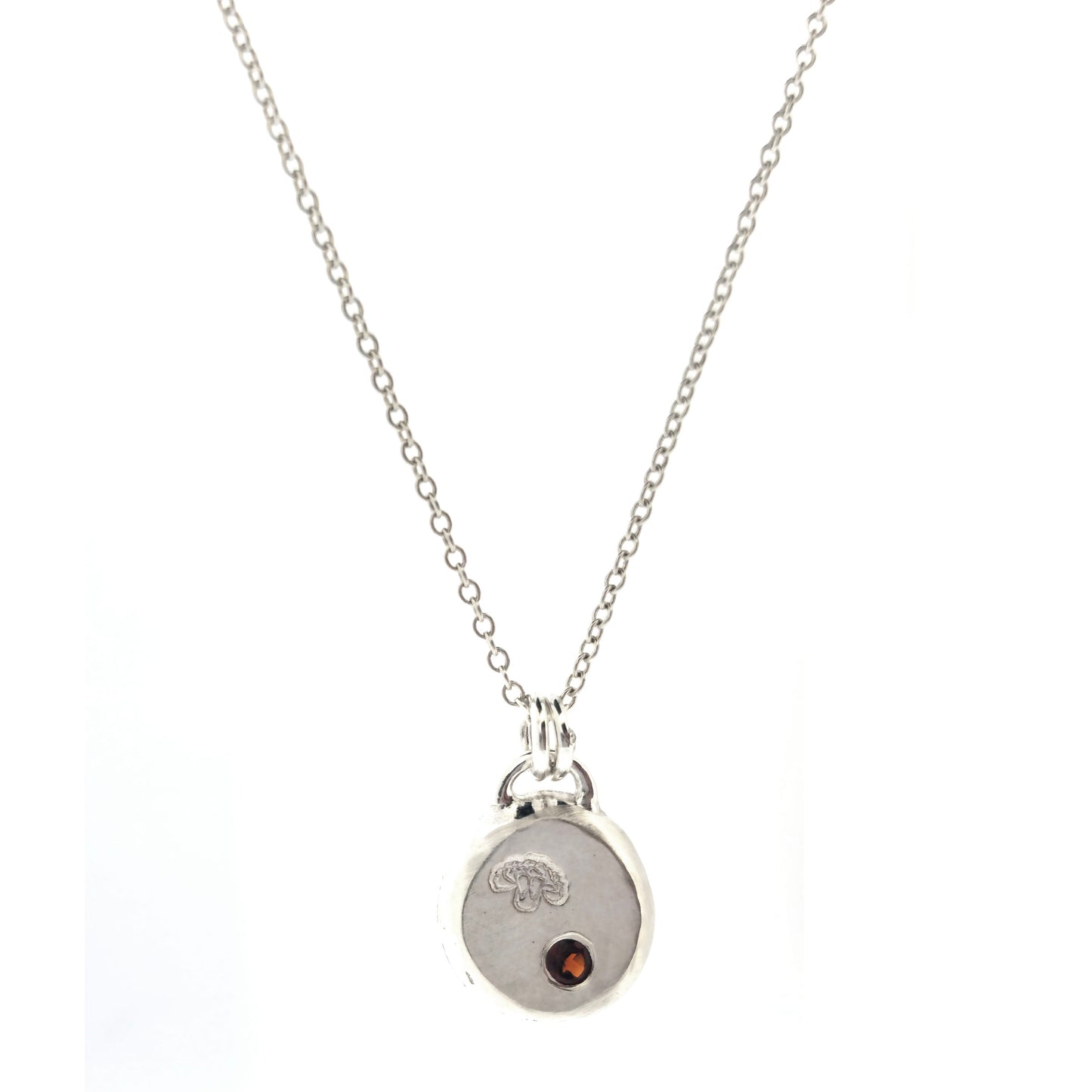 A round silver flat pebble pendant with a carnation flower (January's birth flower) engraved on it and a red garnet gemstone (January's birthstone) flush set into it. On a silver chain. On a white background. Mijoux Jewellery.