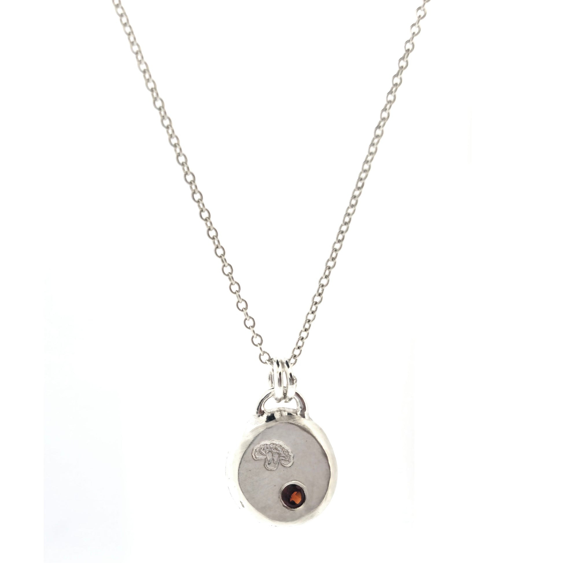 A round silver flat pebble pendant with a carnation flower (January's birth flower) engraved on it and a red garnet gemstone (January's birthstone) flush set into it. On a silver chain. On a white background. Mijoux Jewellery.