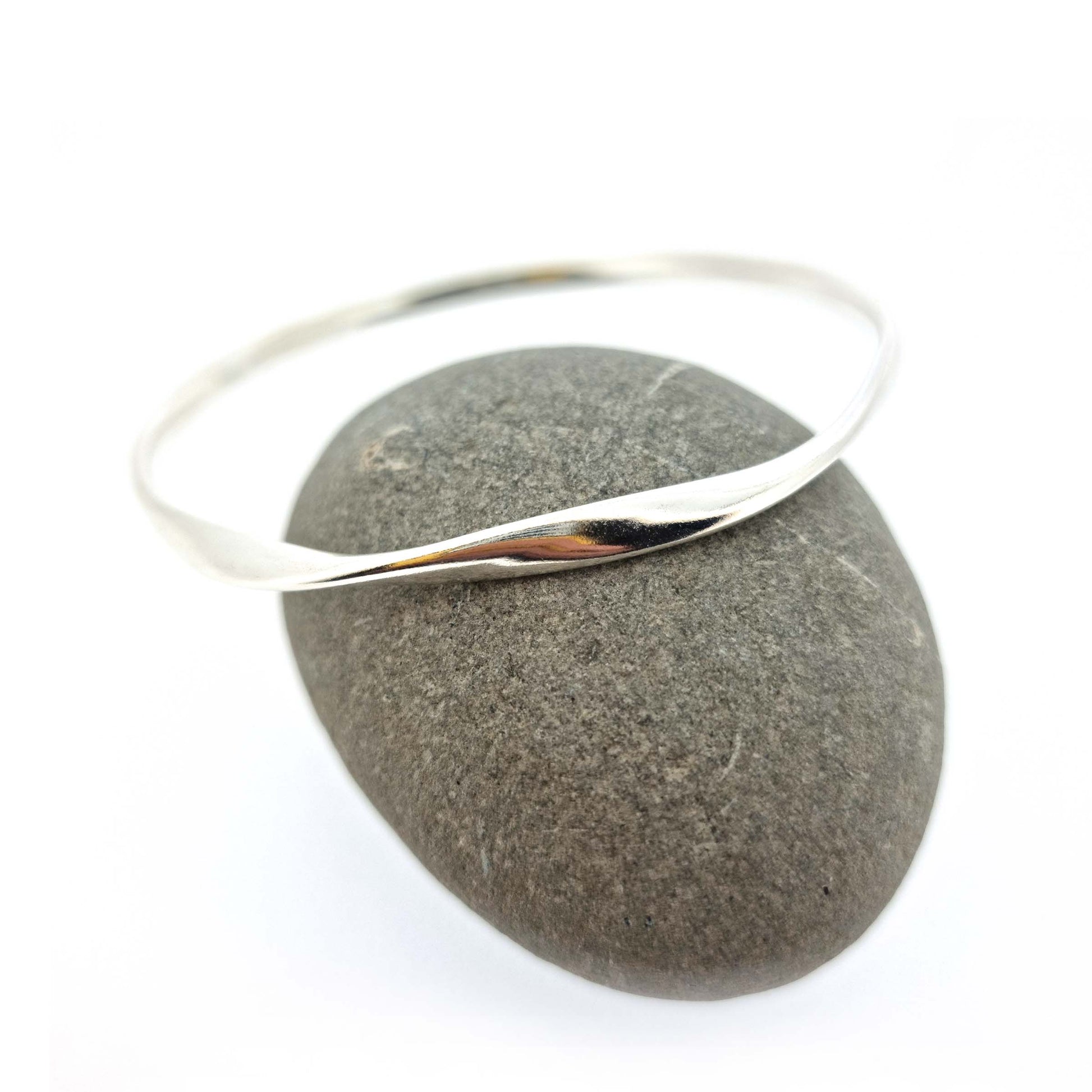 Round silver bangle with oval profile and a twist in one part. Pictured leaning on a pebble.