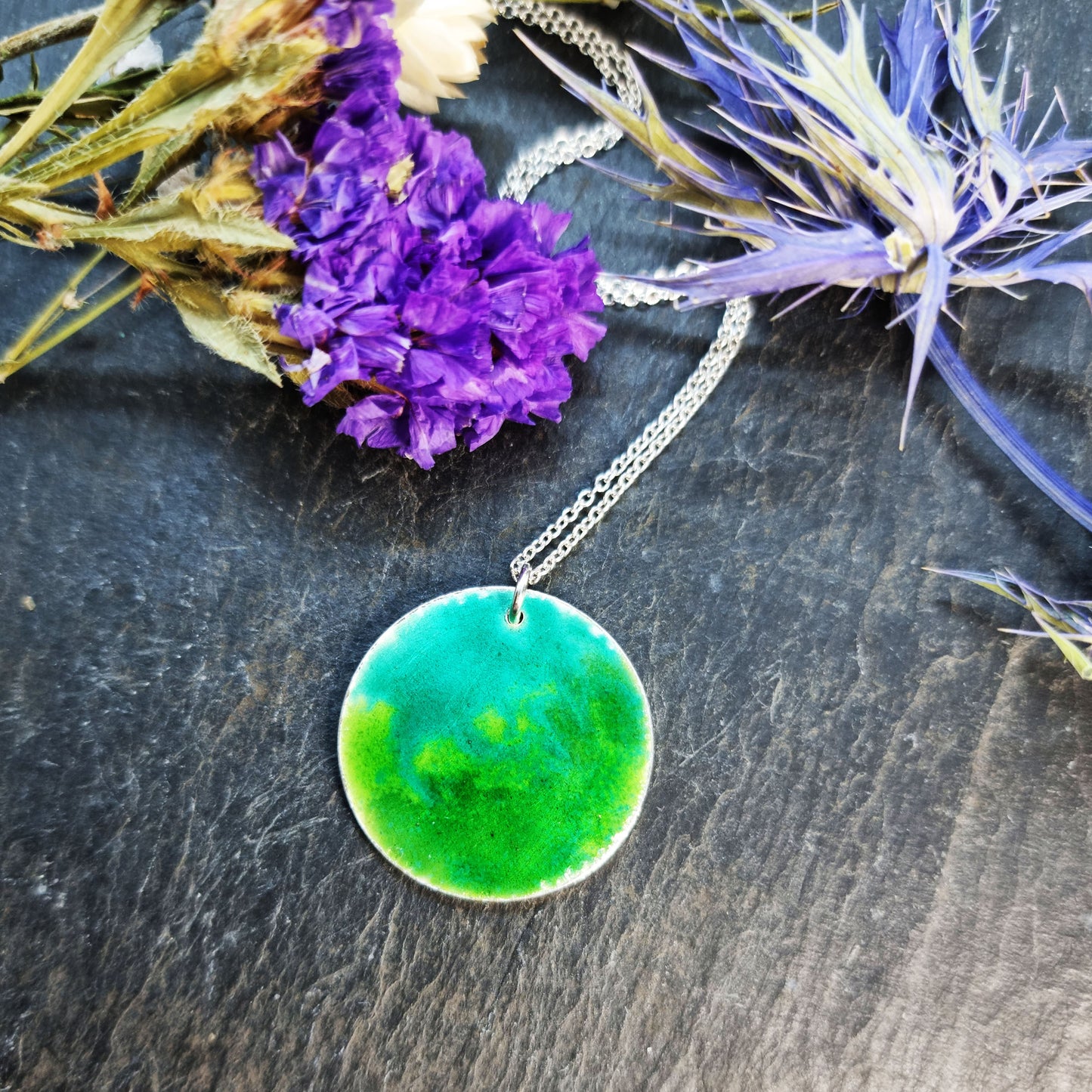 A round silver pendant with a mix of green enamels. on a silver chain. Pictured with flowers.