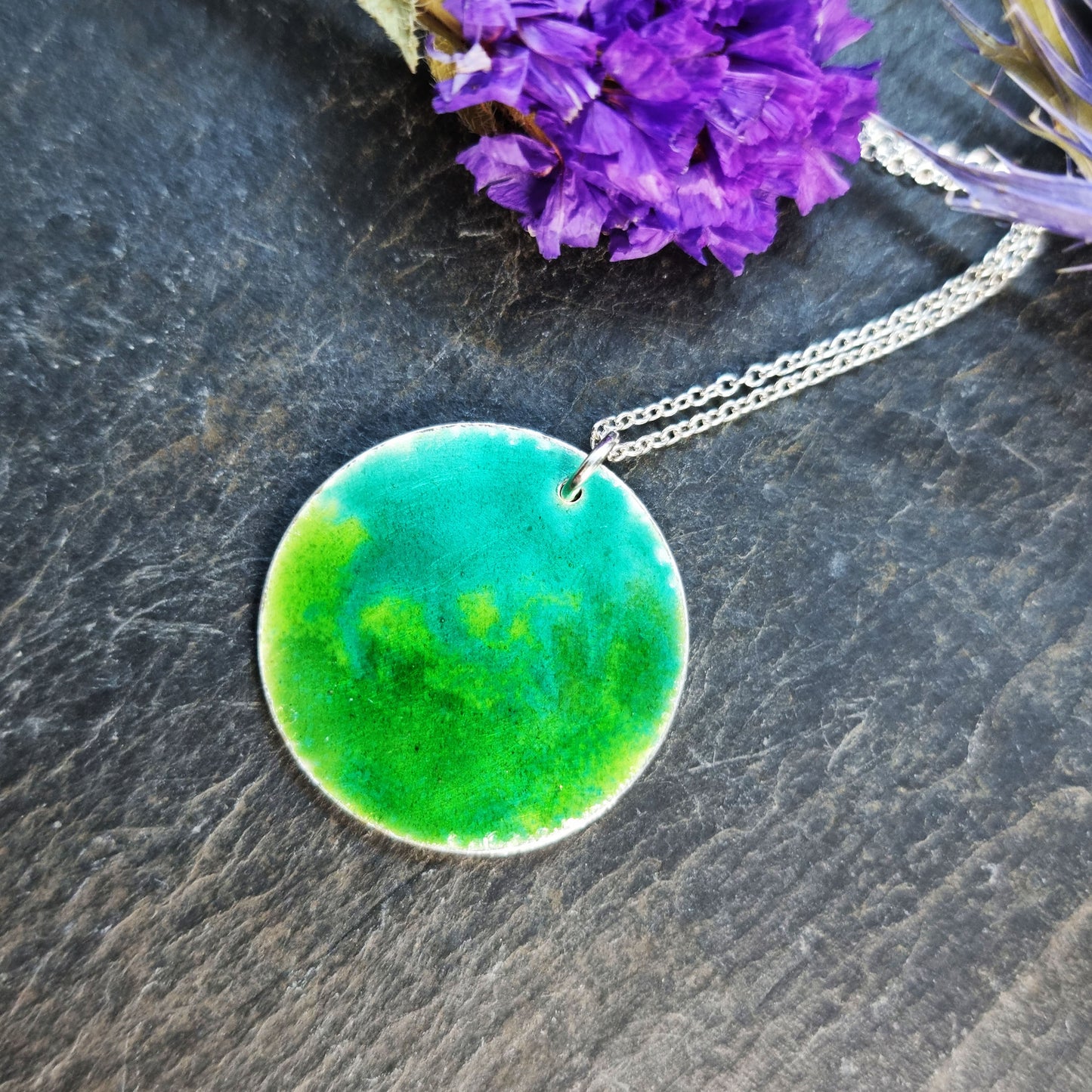 A round silver pendant with a mix of green enamels. on a silver chain. Pictured with flowers.