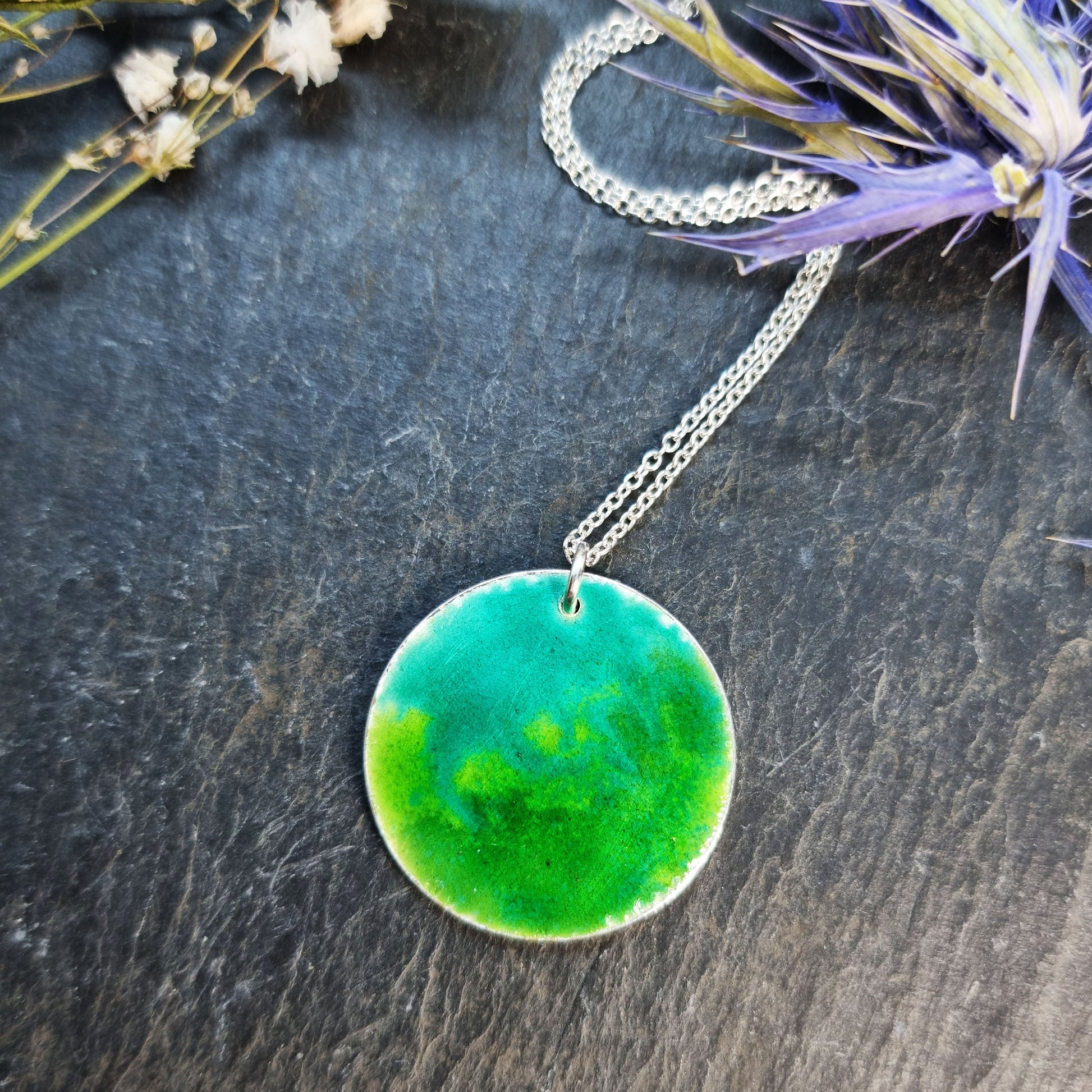 A round silver pendant with a mix of green enamels. on a silver chain. Pictured with flowers.