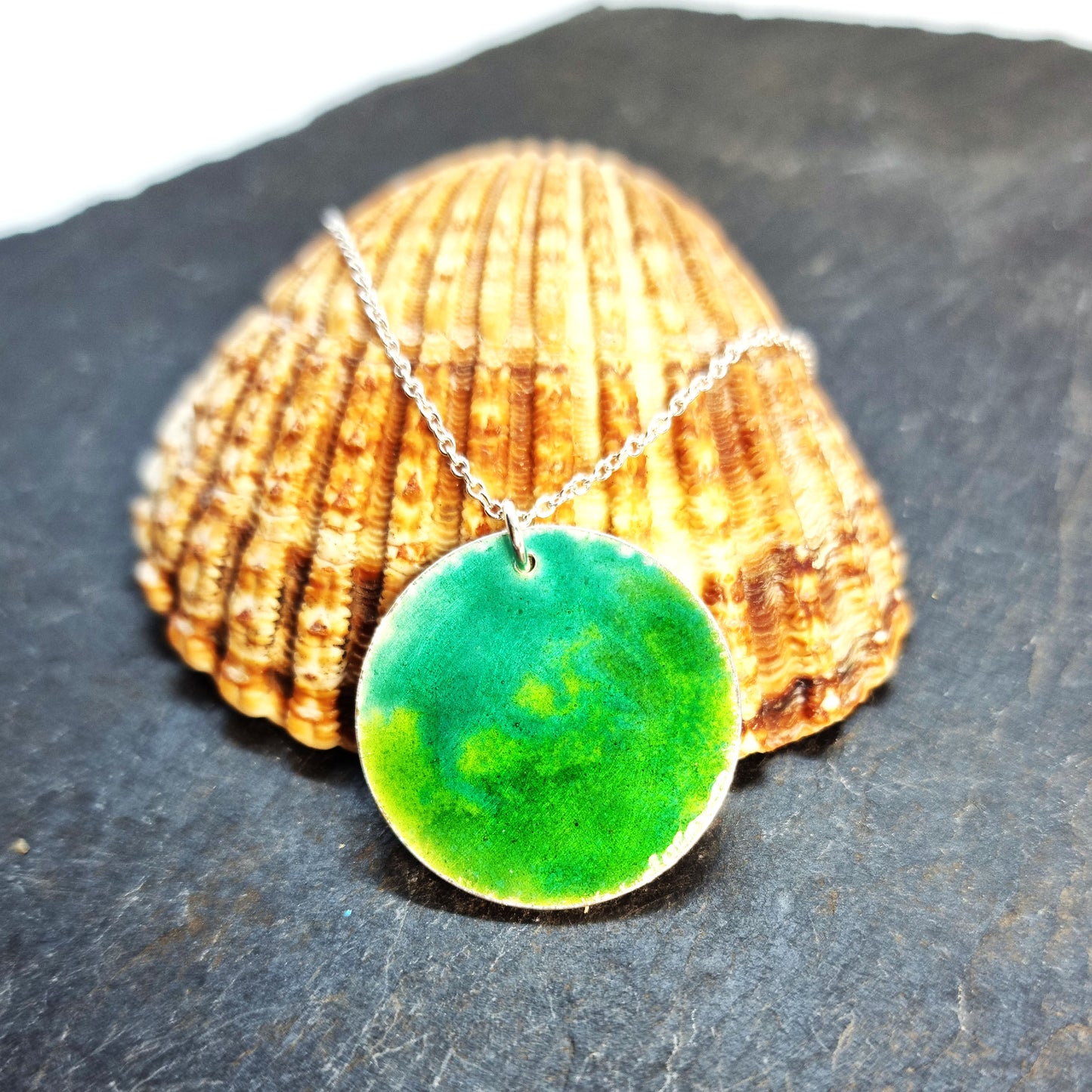 A round silver pendant with a mix of green enamels. on a silver chain. Pictured on a shell.