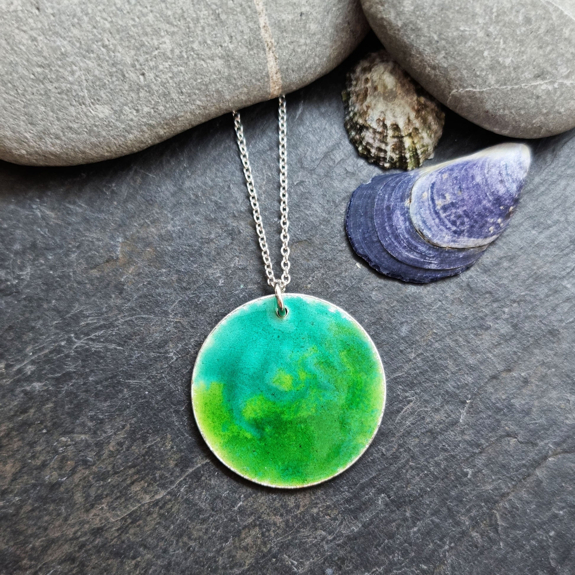 A round silver pendant with a mix of green enamels. On a silver chain. Pictured with pebbles and shells.