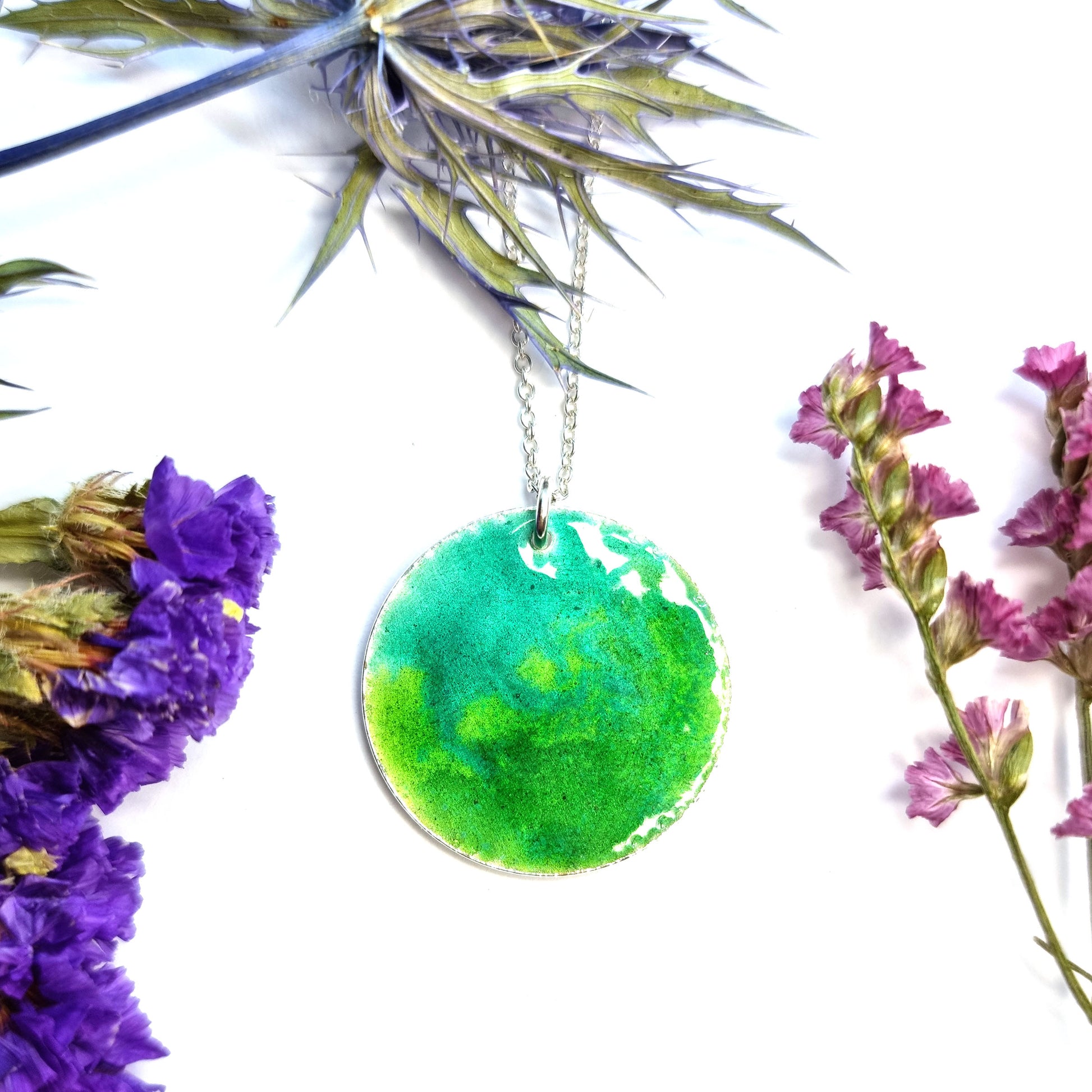 A round silver pendant with a mix of green enamels. on a silver chain. Pictured with flowers.