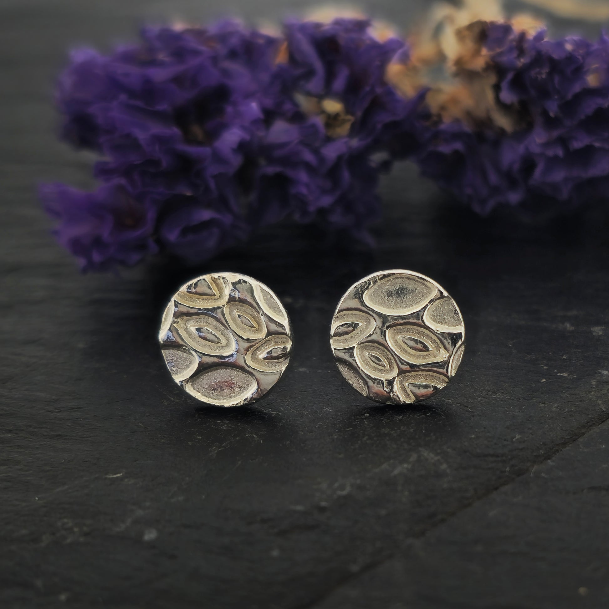 A pair of round silver stud earrings with a marquise (or leaf) shaped pattern on. Shown on slate with flowers.