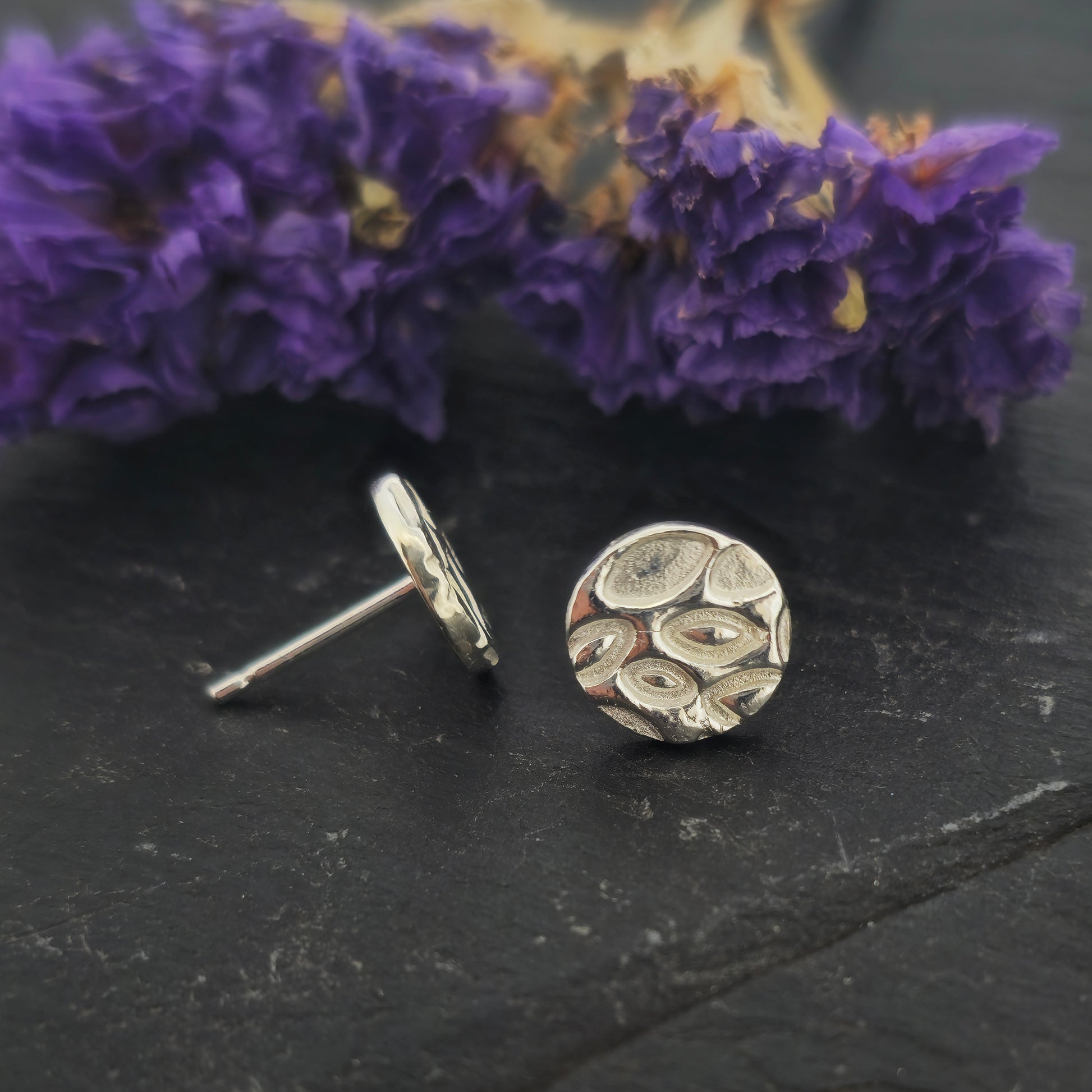 A pair of round silver stud earrings with a marquise (or leaf) shaped pattern on. Shown on slate with flowers. One shows the front and one the side.