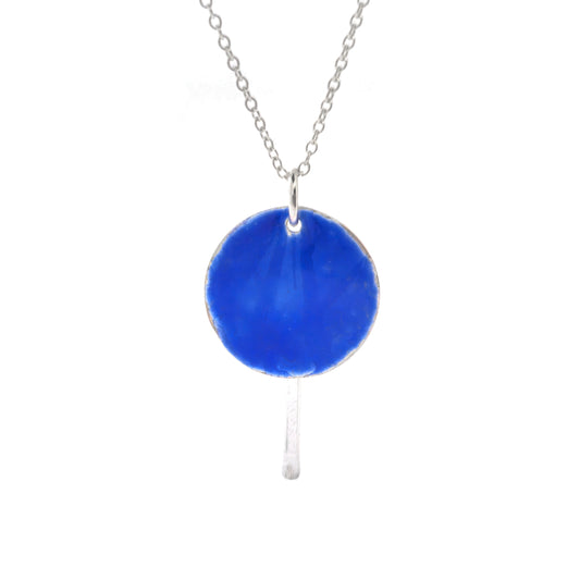 A silver pendant with a round disc that has a lined pattern covered in royal blue enamel. A silver long strip is suspended behind and comes own below the disc. On a silver chain.