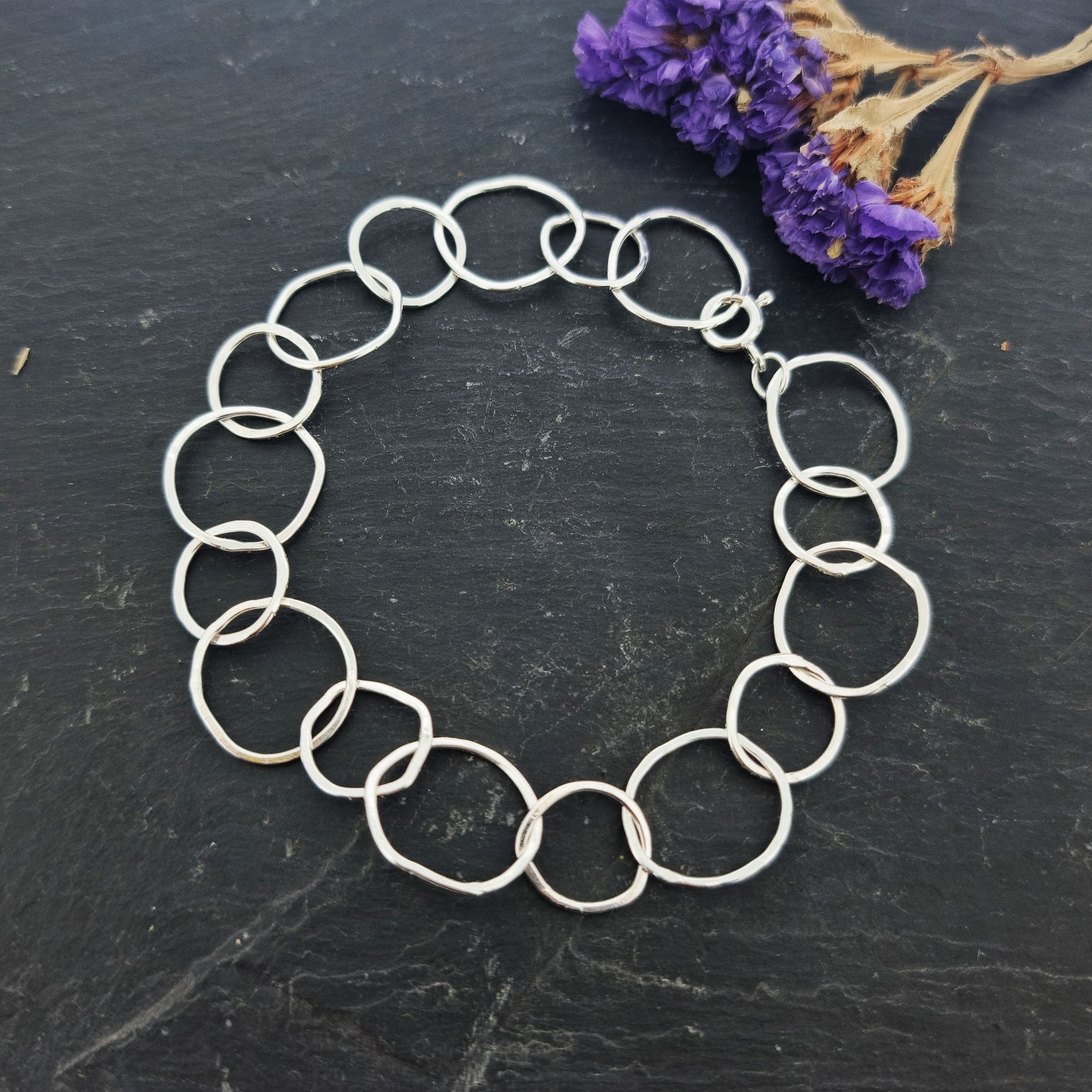 A silver chain link bracelet with delicate links of various sizes in an organic circle shape. Pictured on slate with flowers.