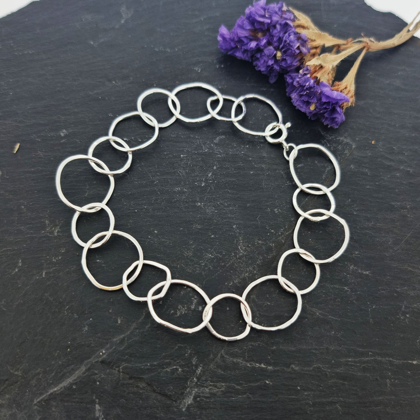 A silver chain link bracelet with delicate links of various sizes in an organic circle shape. Pictured on slate with flowers.