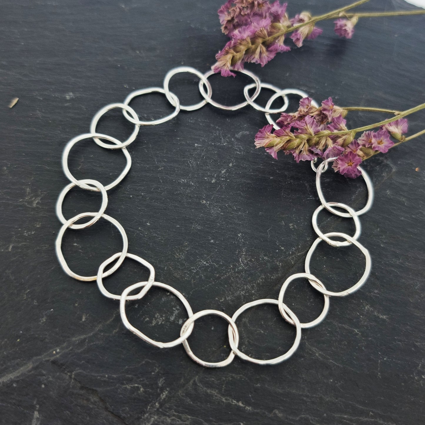 A silver chain link bracelet with delicate links of various sizes in an organic circle shape. Pictured on slate with flowers.