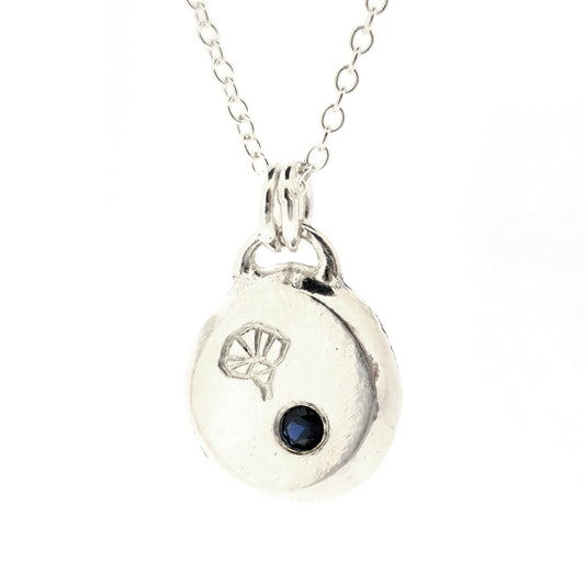 A handmade silver pebble pendant necklace featuring a disc-shaped charm with a small dark blue gemstone and an engraved morning glory flower design, suspended from a delicate silver chain against a white background. Mijoux Jewellery.